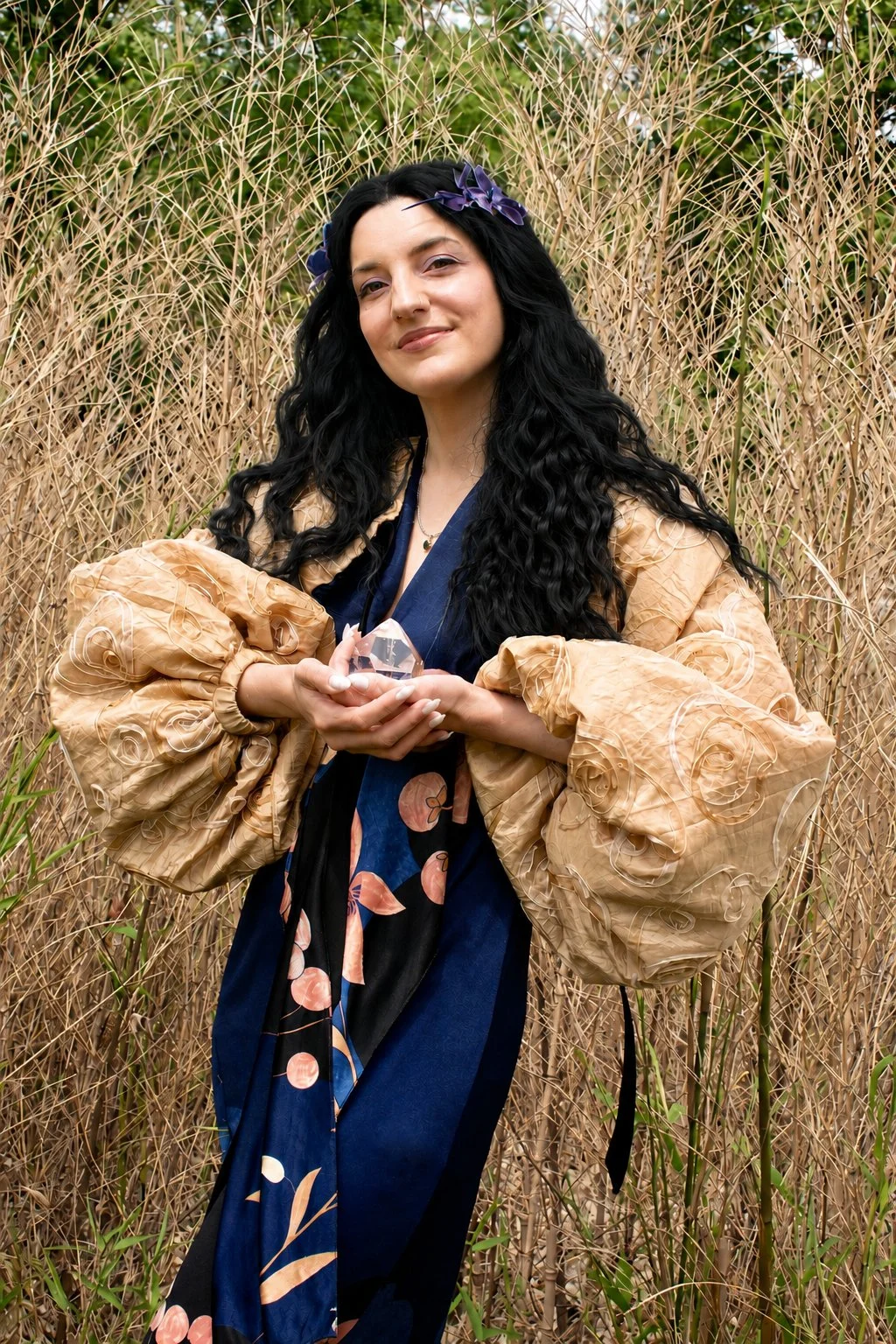 A woman with long, curly black hair and a purple flower headband, dressed in a beige, puff-sleeved jacket over a dark blue dress with floral accents, holding a large clear crystal, standing amidst tall, dried grasses in a natural outdoor setting.