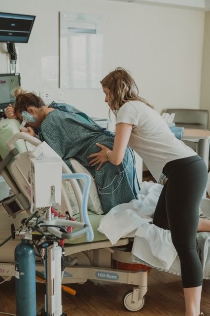 A woman in a white t-shirt and black leggings leaning over a hospital bed, holding a patient's hand and providing support in a hospital room.