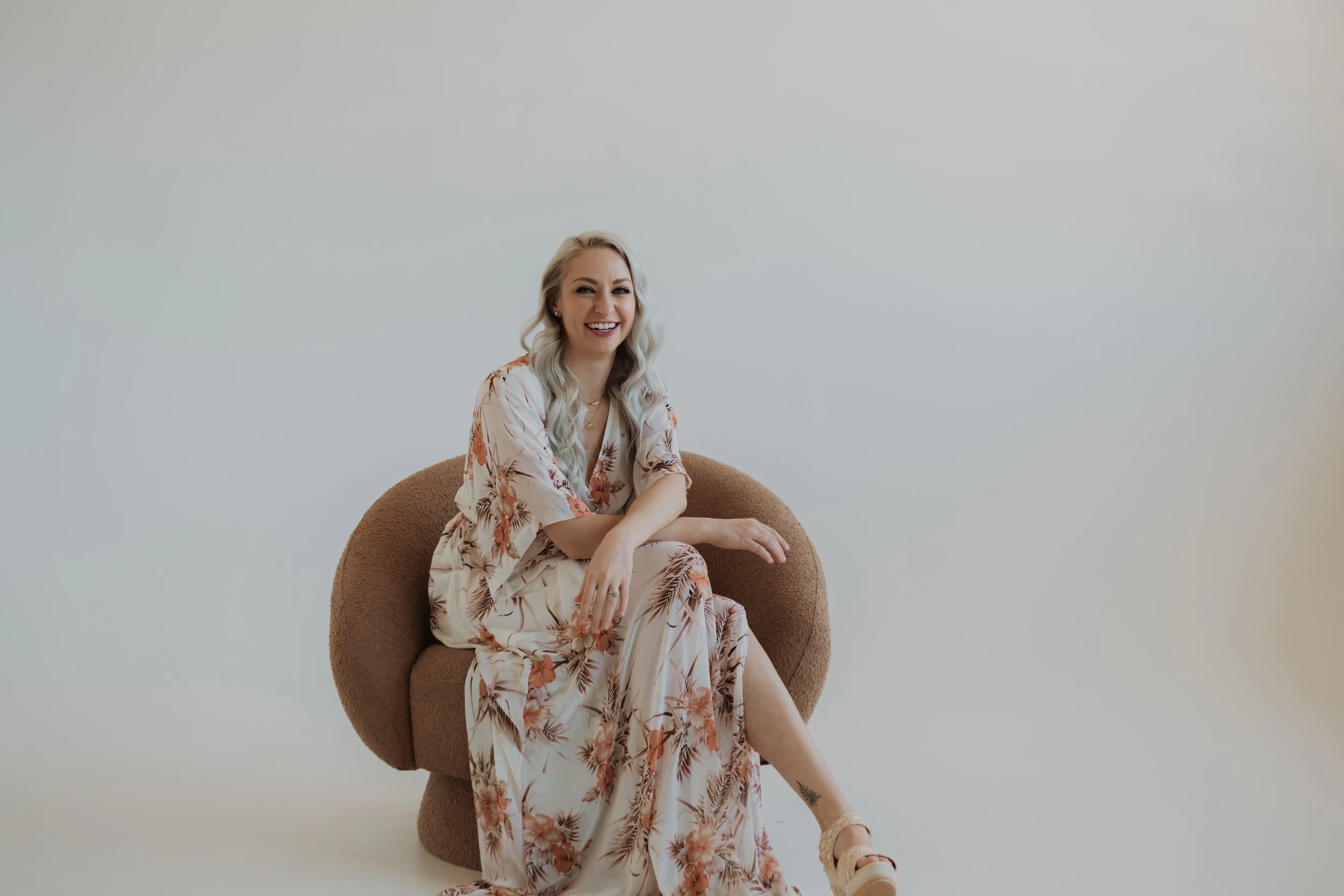A woman sitting on a plush, brown armchair against a plain, light-colored wall, smiling at the camera. She has long, wavy blonde hair and is wearing a flowing, floral-patterned dress.