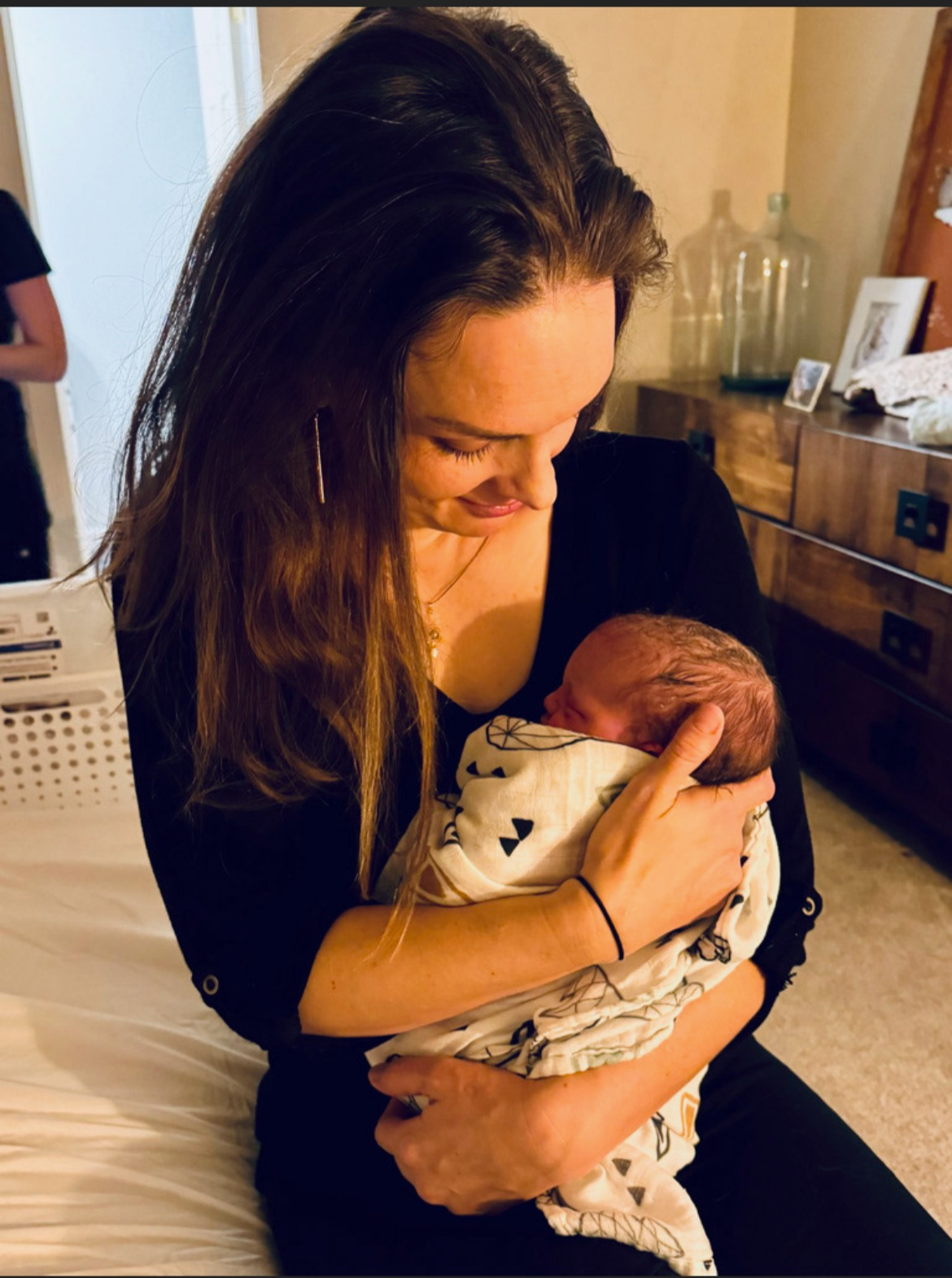 Woman holding a newborn baby in a warm, intimate moment inside a room with wooden furniture and decorative items.