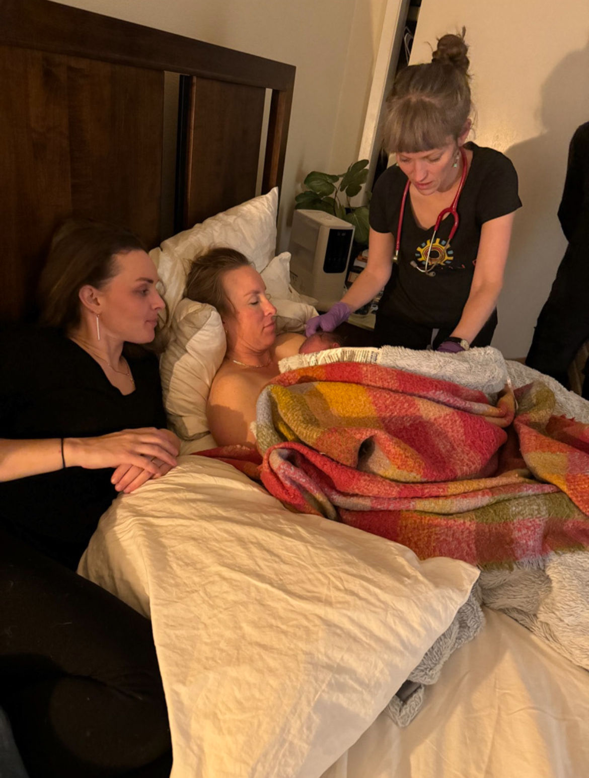 A woman giving birth in a hospital bed with two women beside her, a healthcare worker with a stethoscope attending to the newborn, and a blanket covering the mother.