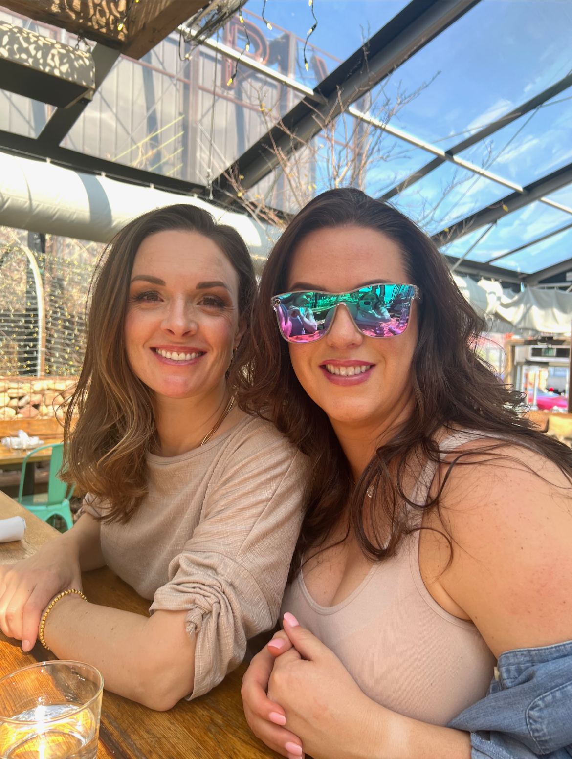 Two women smiling at a restaurant or cafe with a glass roof and outdoor seating, one wearing sunglasses with reflective lenses, and the other with wavy hair and a beige top.