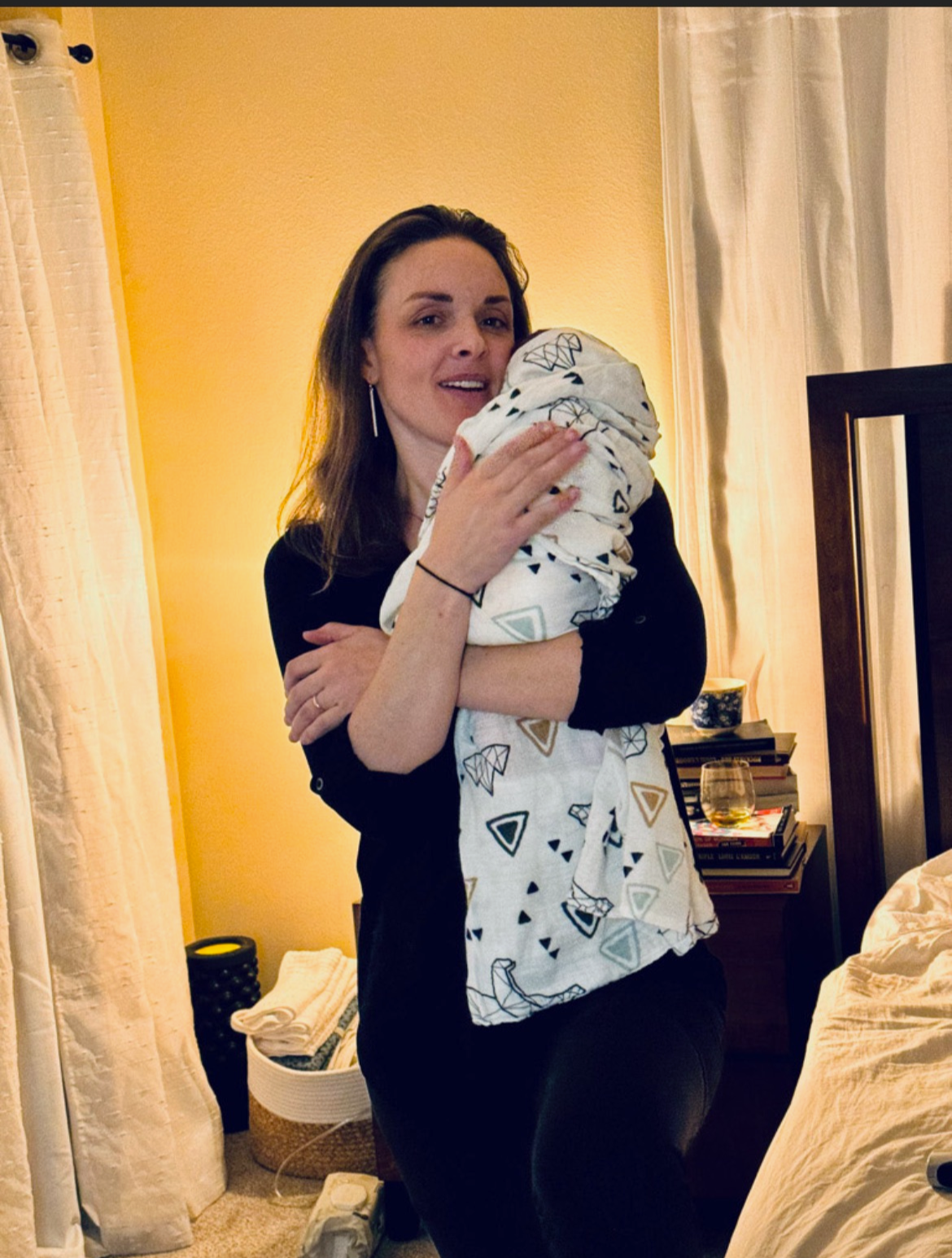 A woman with shoulder-length brown hair and a black top holding a baby wrapped in a blanket with geometric patterns in a warmly lit bedroom with yellow walls and beige curtains.