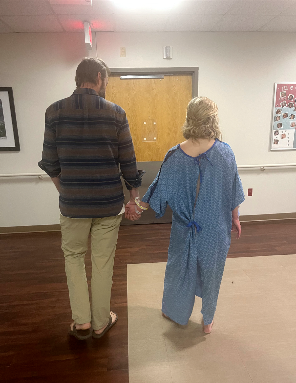 A woman in a hospital gown holding hands with a man wearing casual clothing, walking down a hallway.