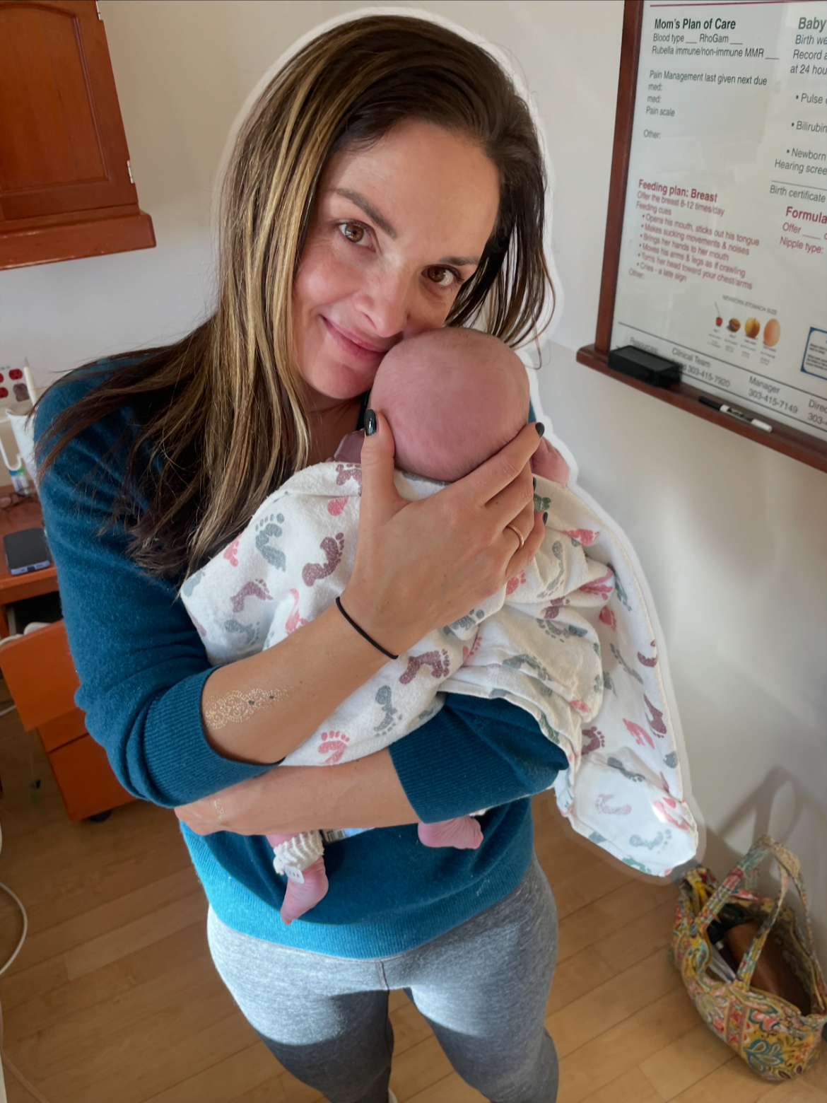 Woman holding a newborn baby in a hospital room, close to a care plan chart on the wall.