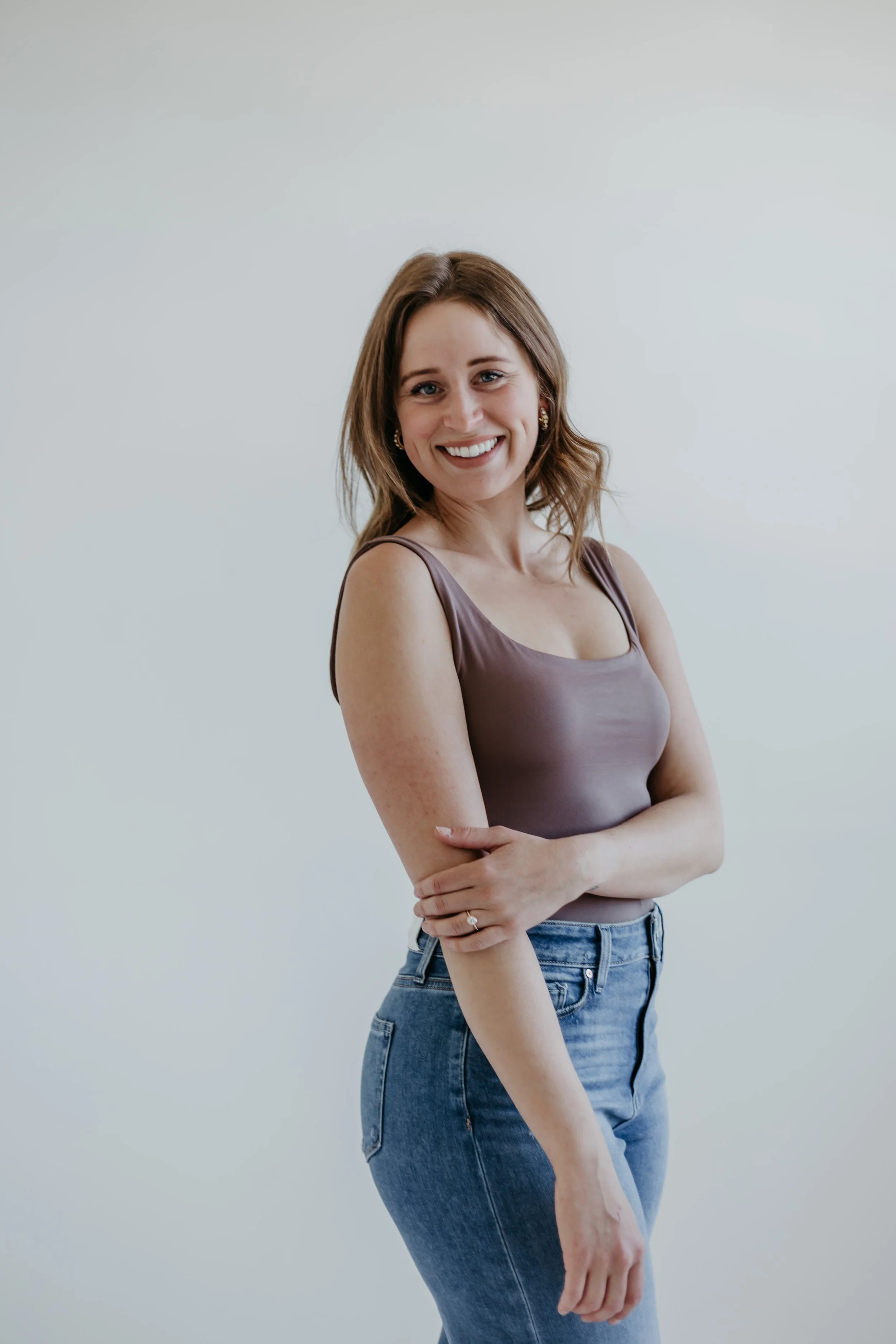 A woman with light brown hair and earrings, smiling, wearing a mauve tank top and blue jeans, standing against a plain, light-colored background.