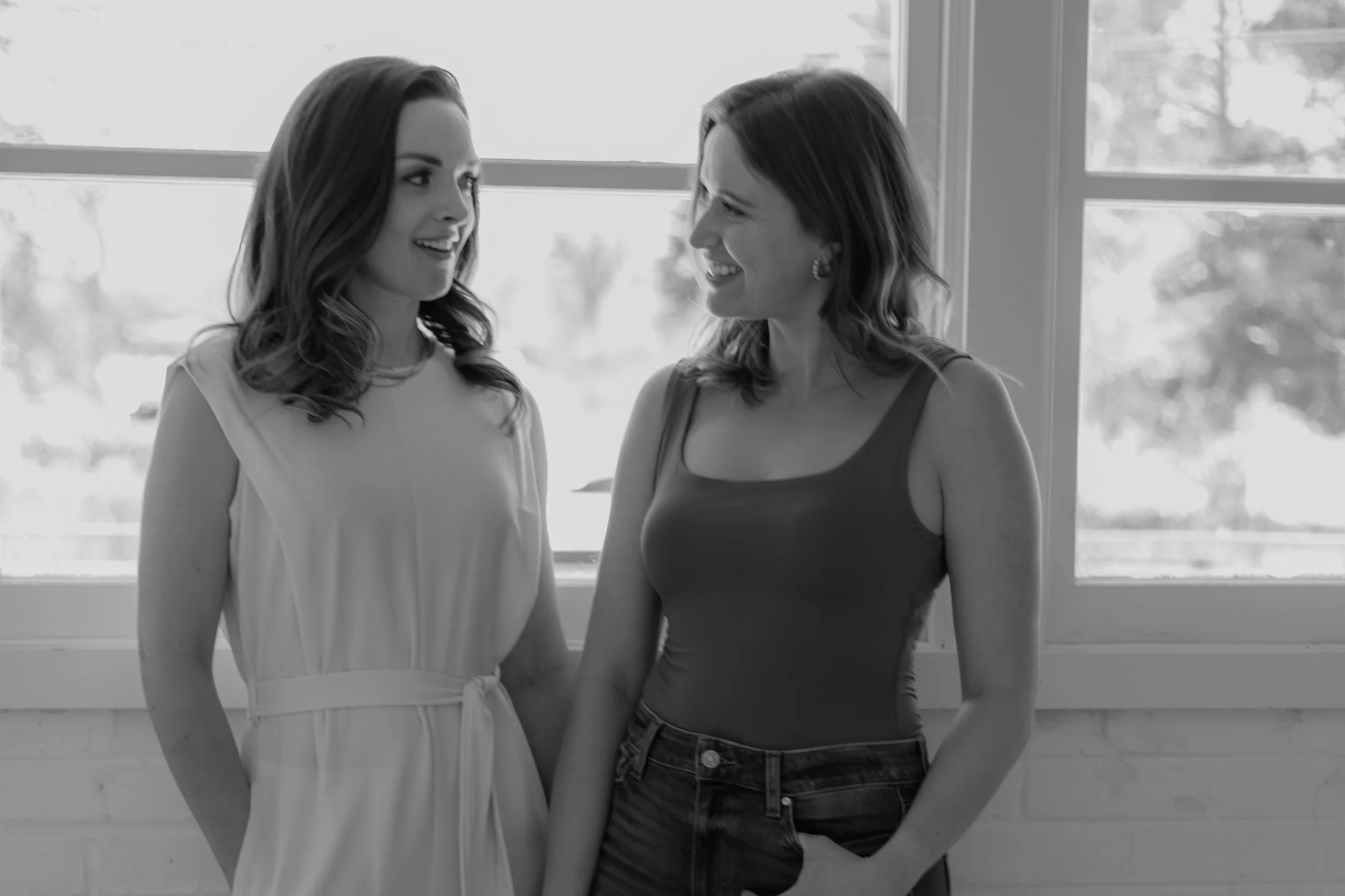 Two women standing close to each other indoors in front of a large window, smiling and looking at each other.