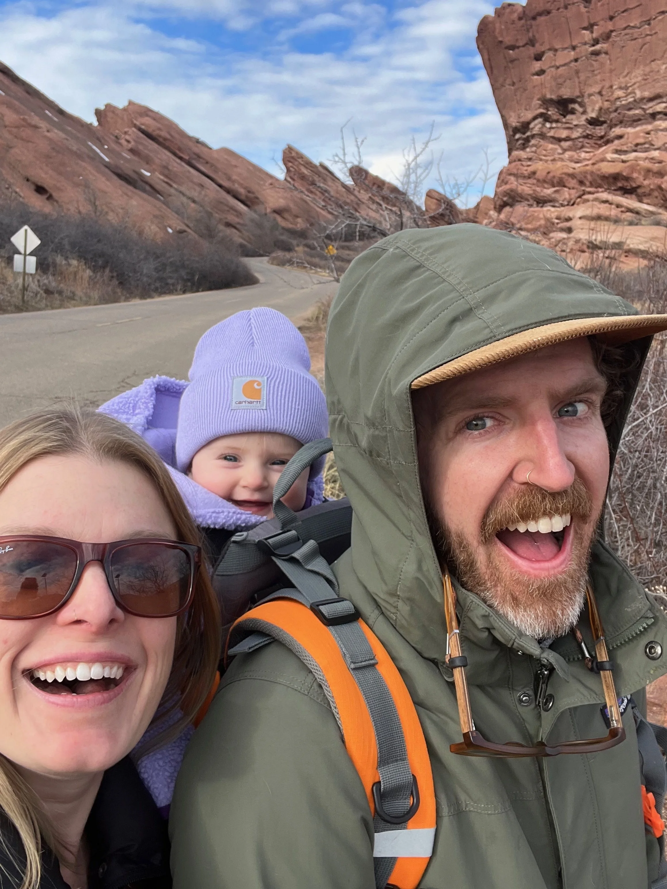 Family of three taking a selfie outdoors with rocks and a cloudy sky in the background. The man is wearing a green hooded jacket and has a beard. The woman has brown hair, glasses, and is smiling. The child is wearing a purple beanie and jacket, and 