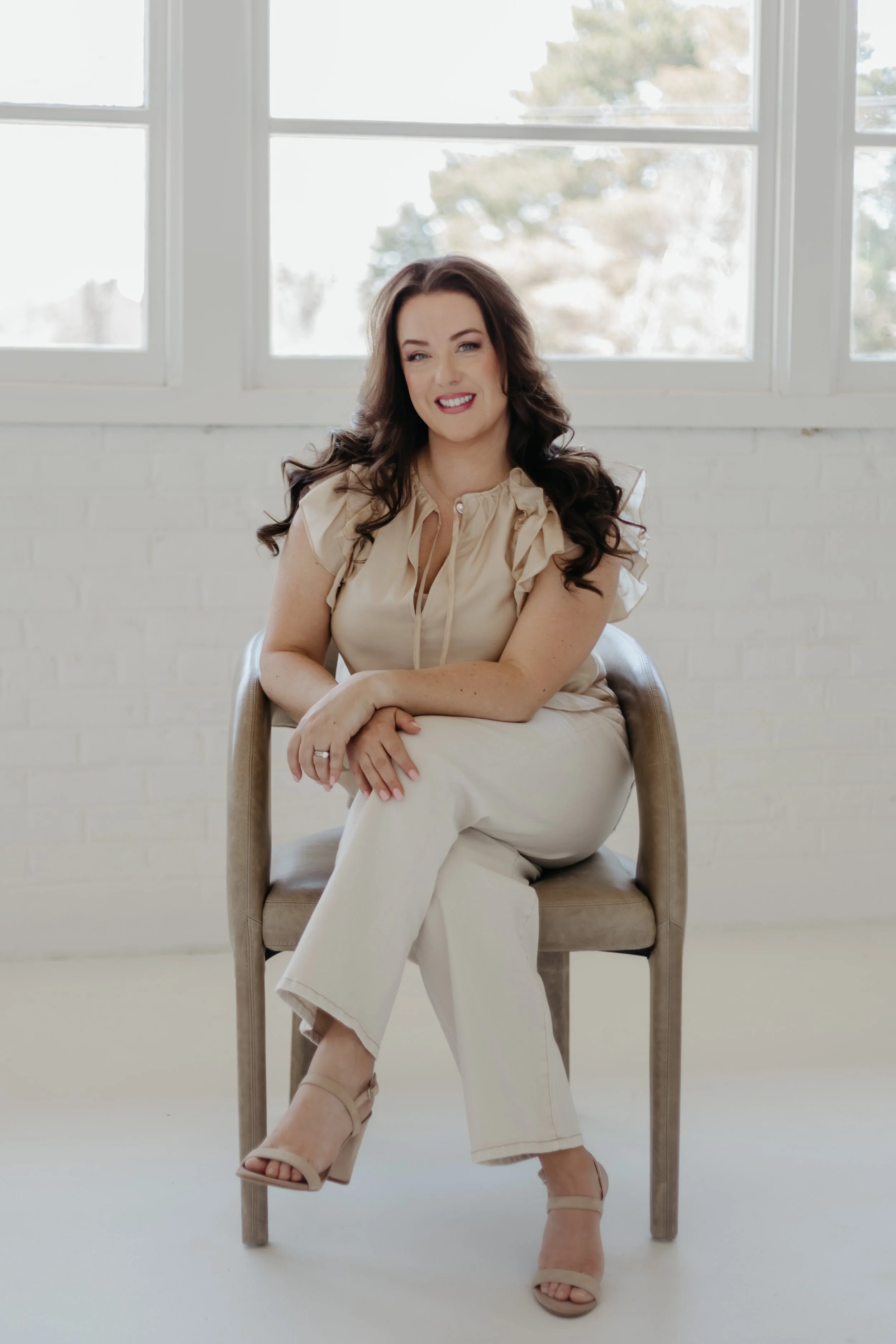 A woman with long dark hair wearing a beige blouse and beige pants sitting on a beige chair in front of large windows with white frames, outside view of trees and sky.