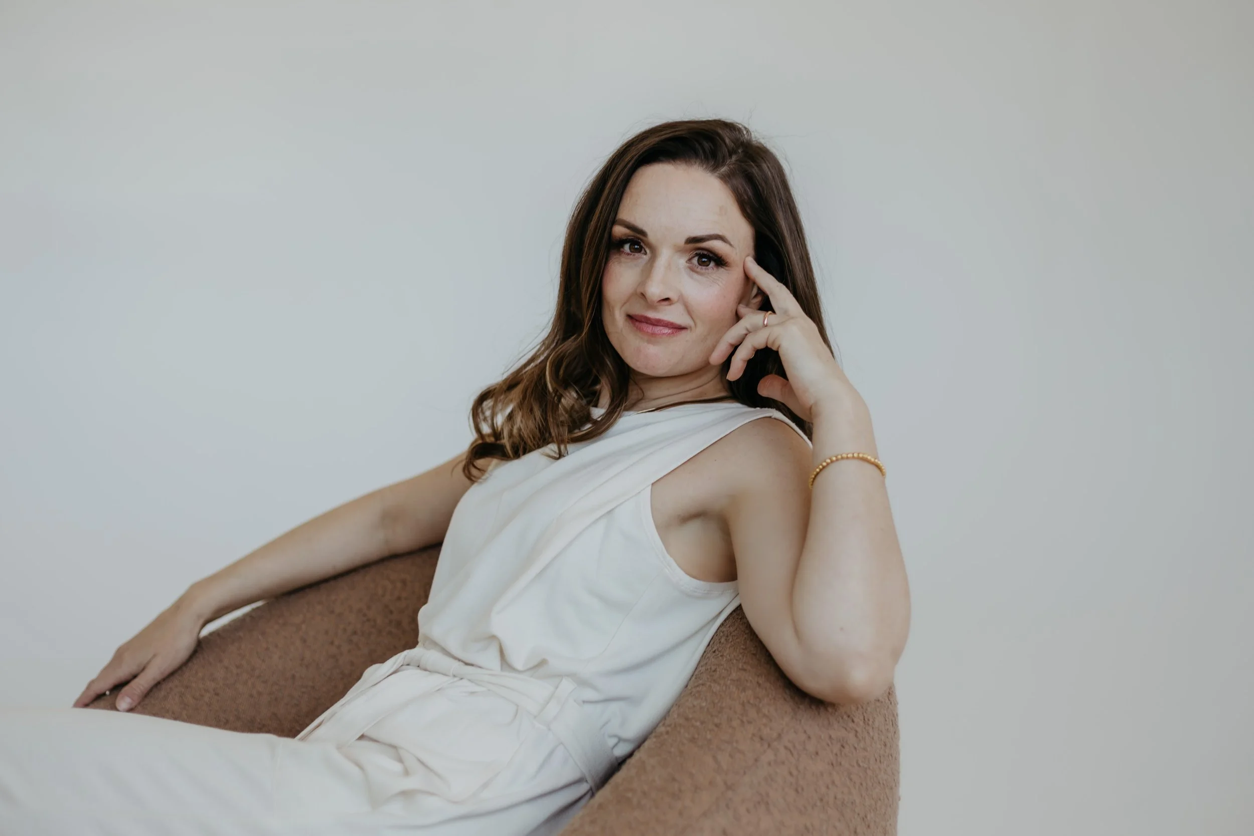 A woman with brown hair wearing a sleeveless white top, sitting on a brown chair, resting her head on her right hand, and smiling gently at the camera.