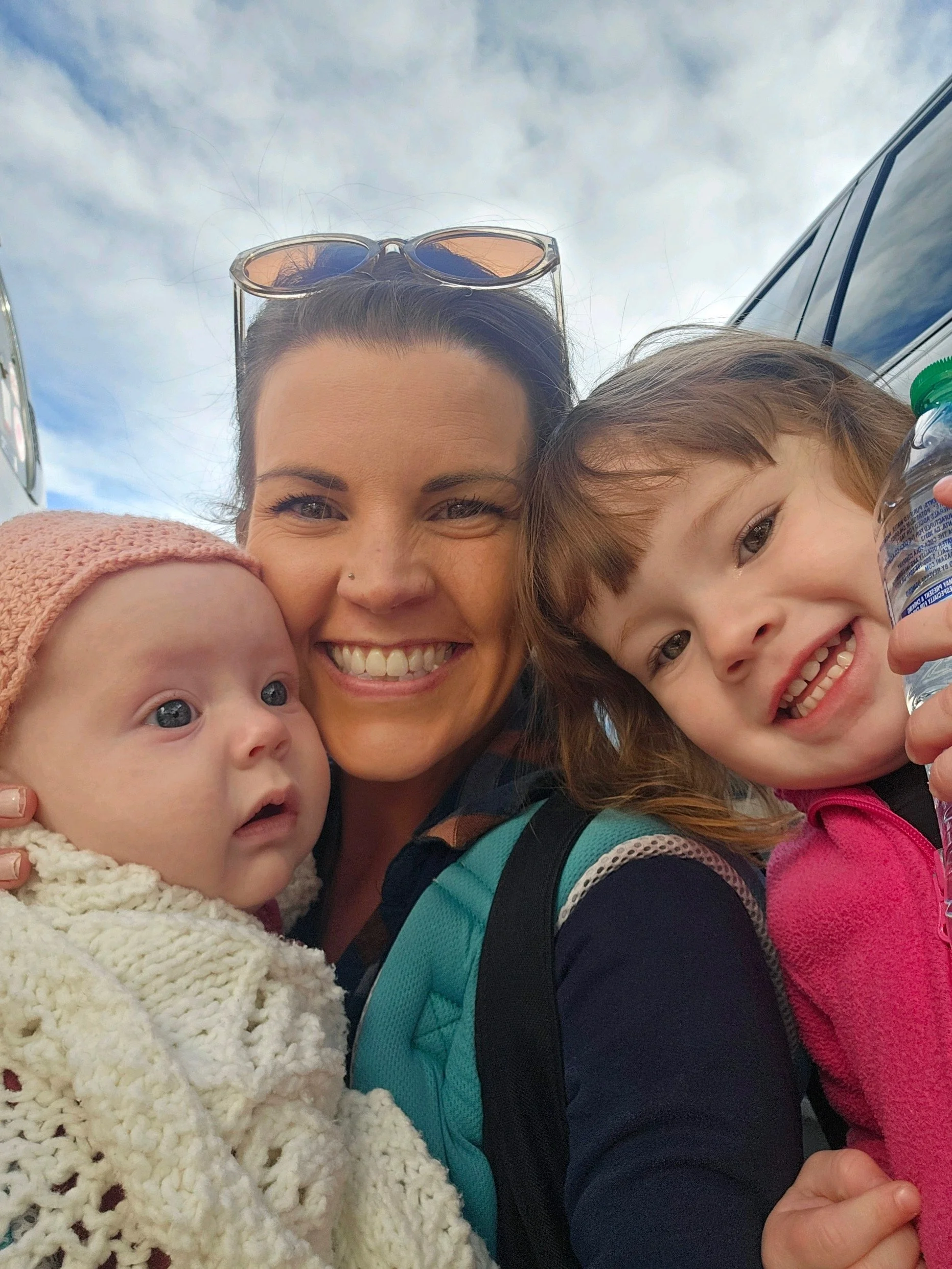 A woman with sunglasses on her head and two children smiling closely together outdoors against a cloudy sky, with a car partly visible in the background.