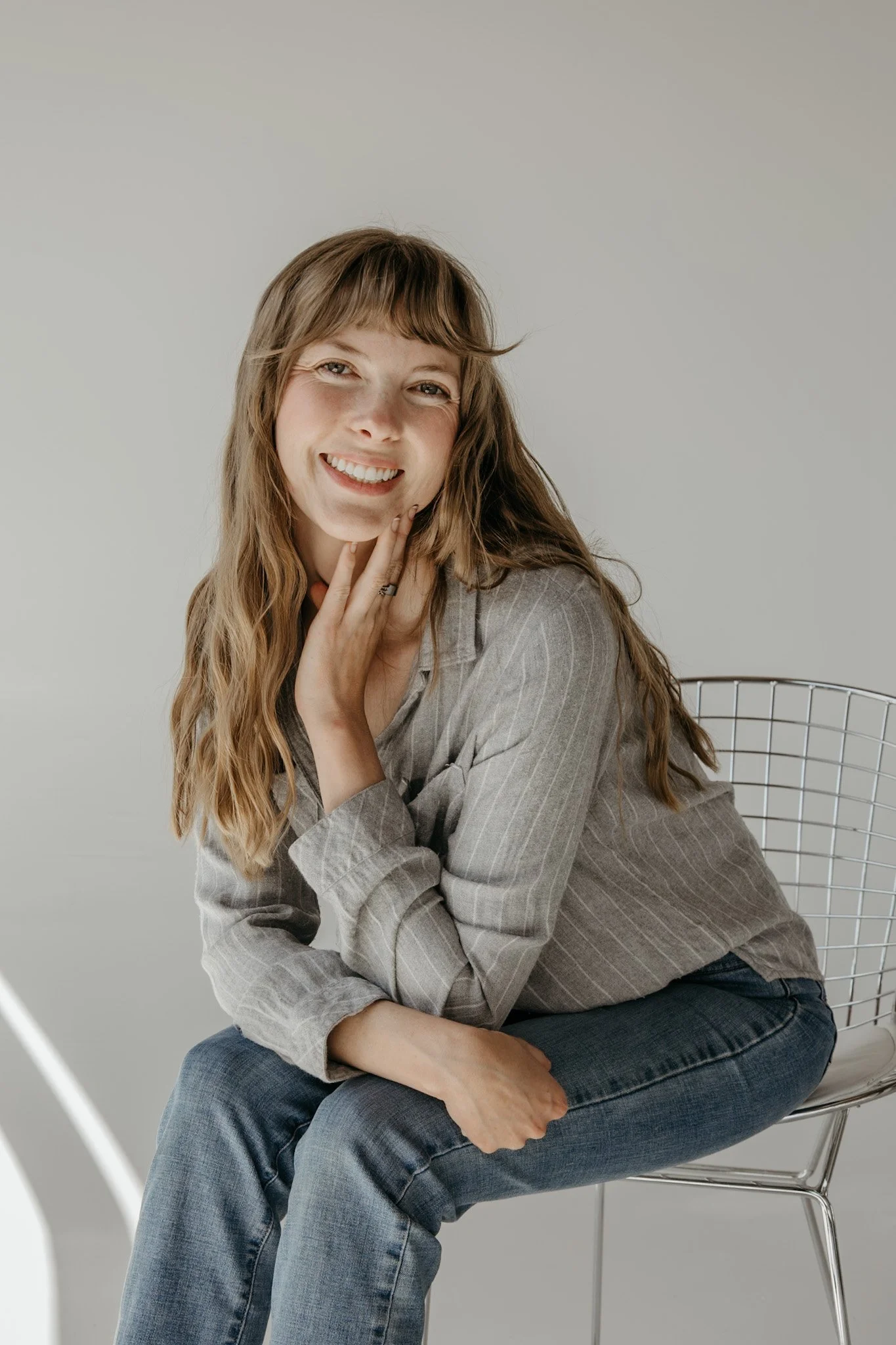 A young woman with long wavy hair, smiling, sitting on a metal wire chair against a plain light gray background. She wears a gray striped button-up shirt and blue jeans.