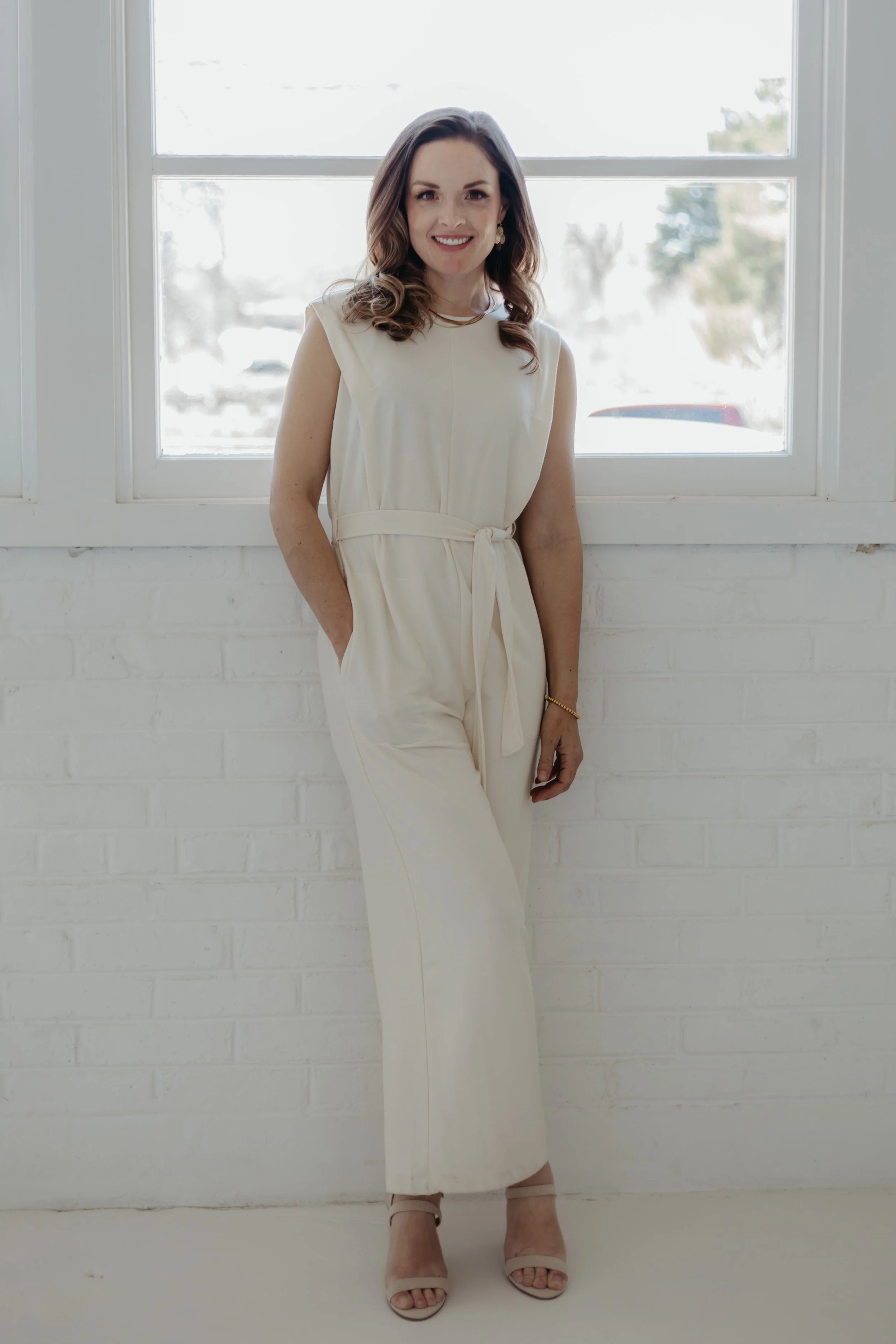 A woman with wavy brown hair wearing an off-white sleeveless jumpsuit, standing in front of a window with a white brick wall, smiling and looking at the camera.