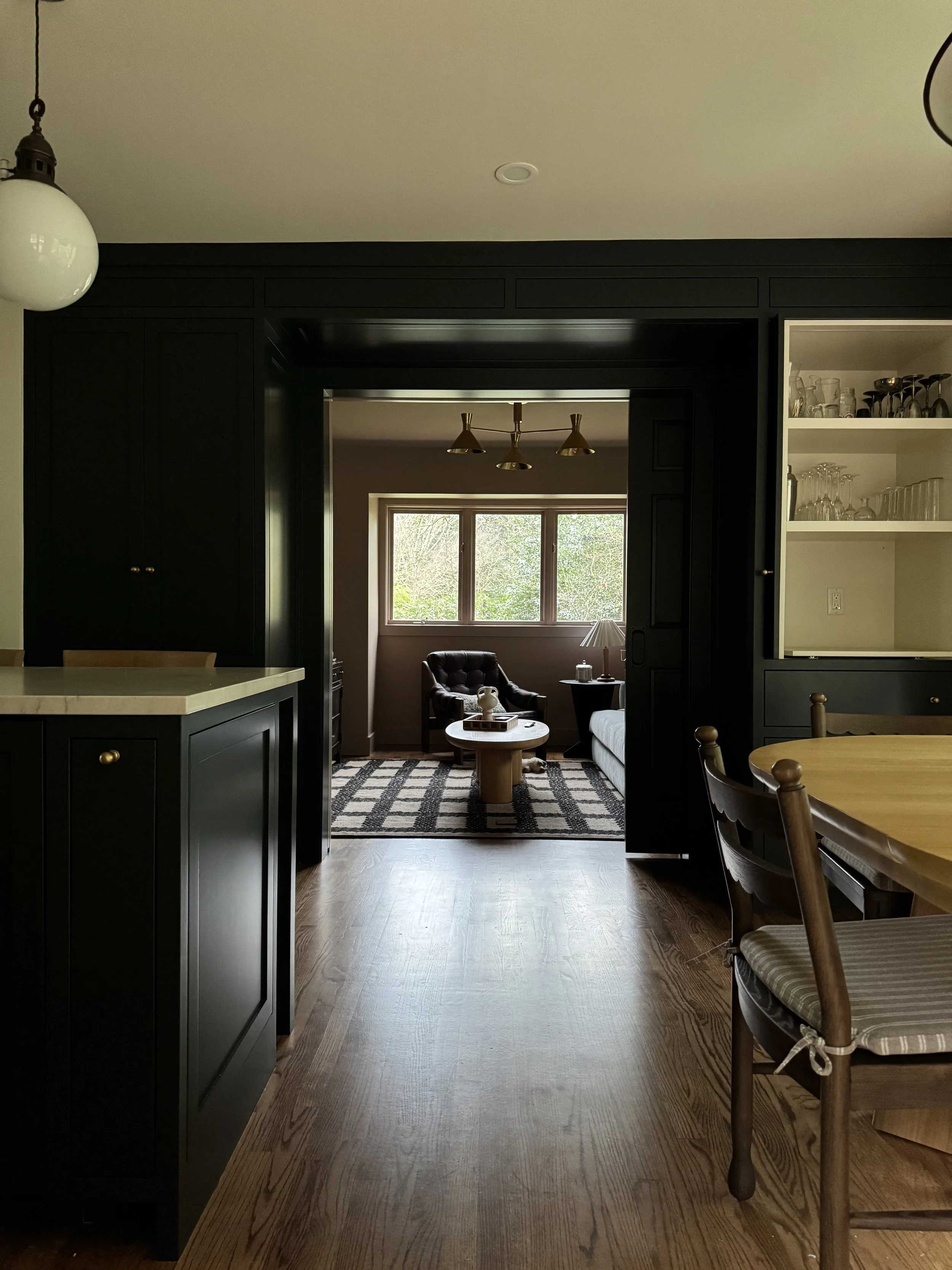 View through a doorway into a cozy living room with a window, armchair, coffee table, and standing lamp, with a dining area to the right and black cabinetry framing the doorway.