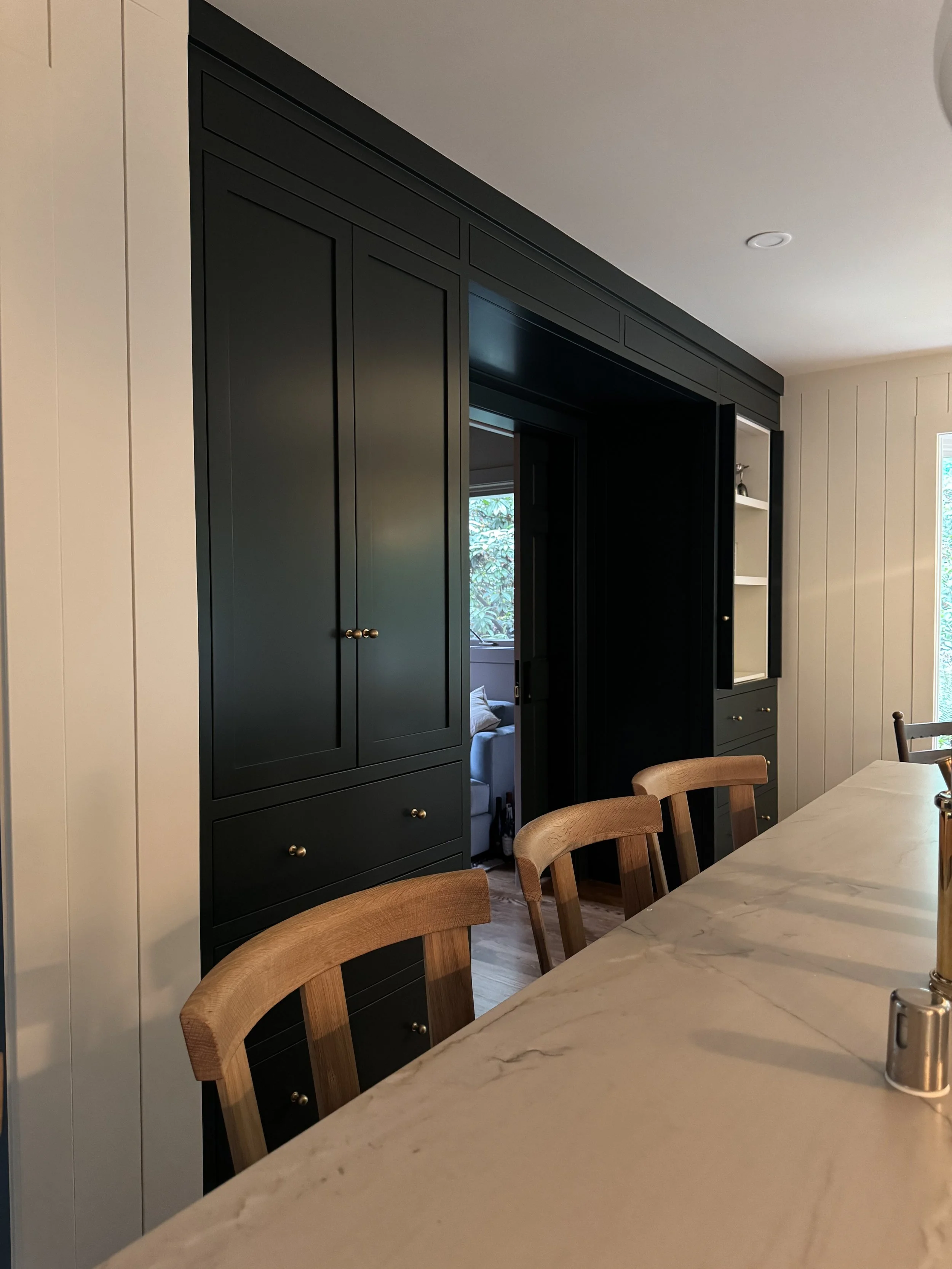 A kitchen or dining area with a large black cabinet and a marble tabletop with wooden chairs. Natural light from a window illuminates the space.