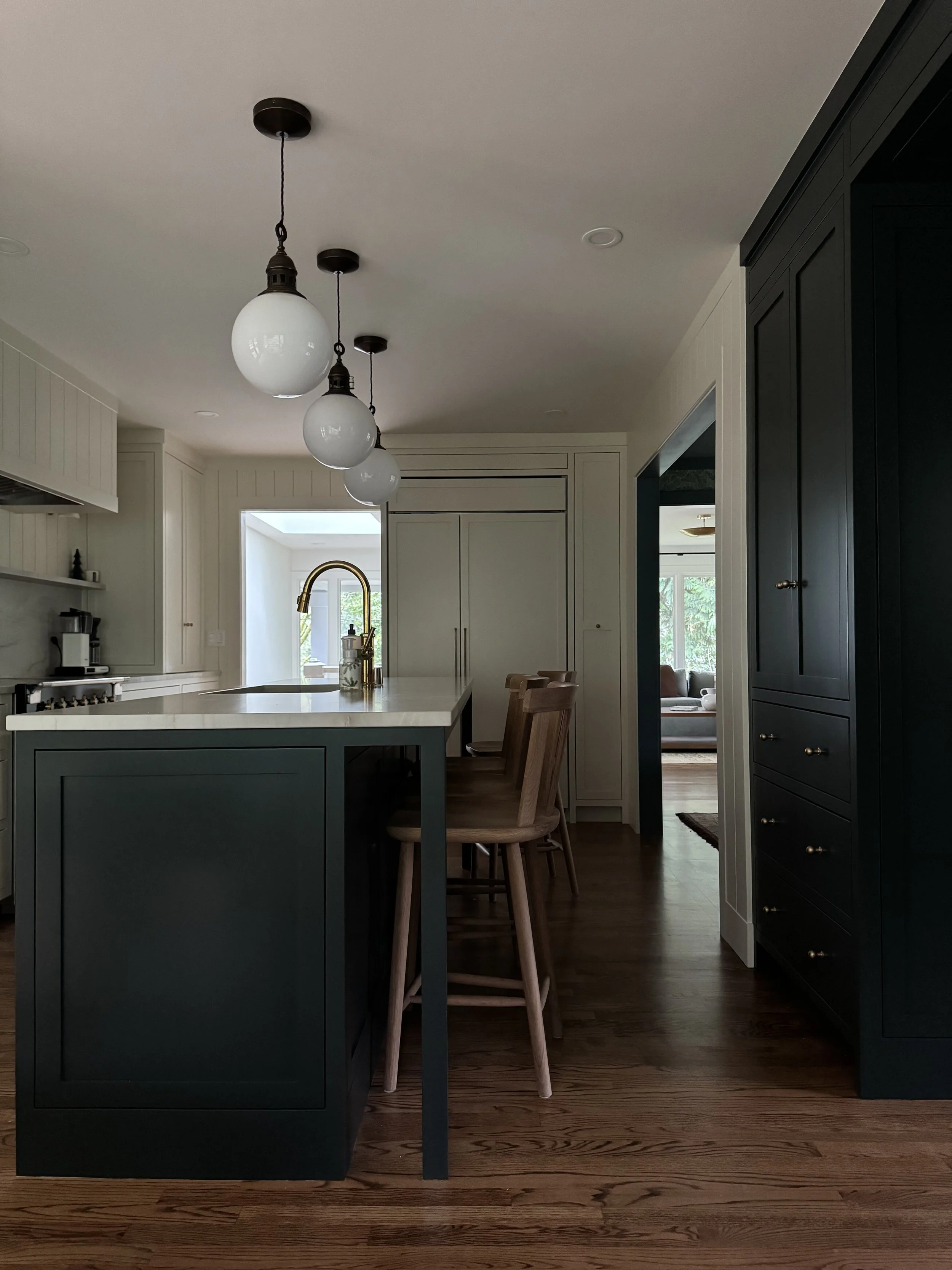 Modern kitchen with dark green island, white countertop, wooden bar stools, overhead white glass pendant lights, and dark cabinetry, with a view into a living room.