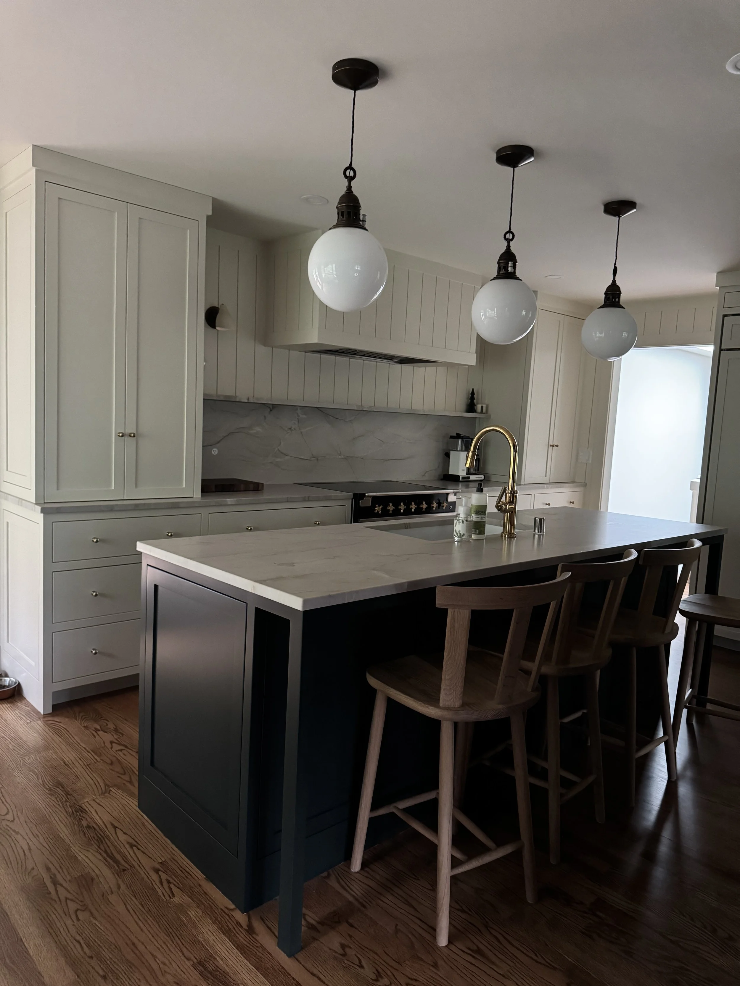Modern kitchen with white cabinets, a marble backsplash, a black island with a white marble countertop, and three hanging pendant lights. There are wooden bar stools at the island and hardwood floors.