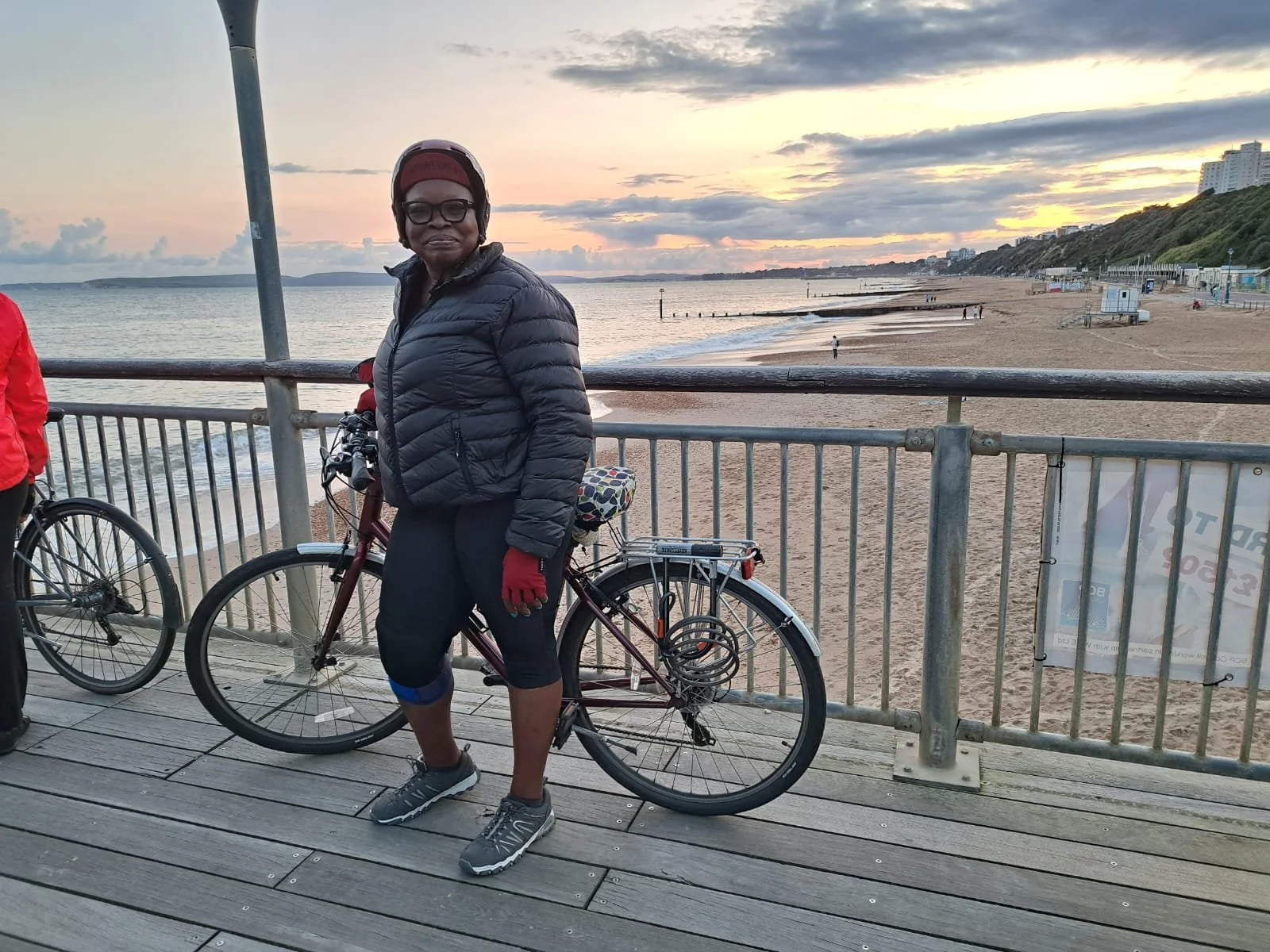 Woman standing with a bicycle on a boardwalk at the beach during sunset, wearing a black puffer jacket, red gloves, black cap, sunglasses, and gray athletic shoes.