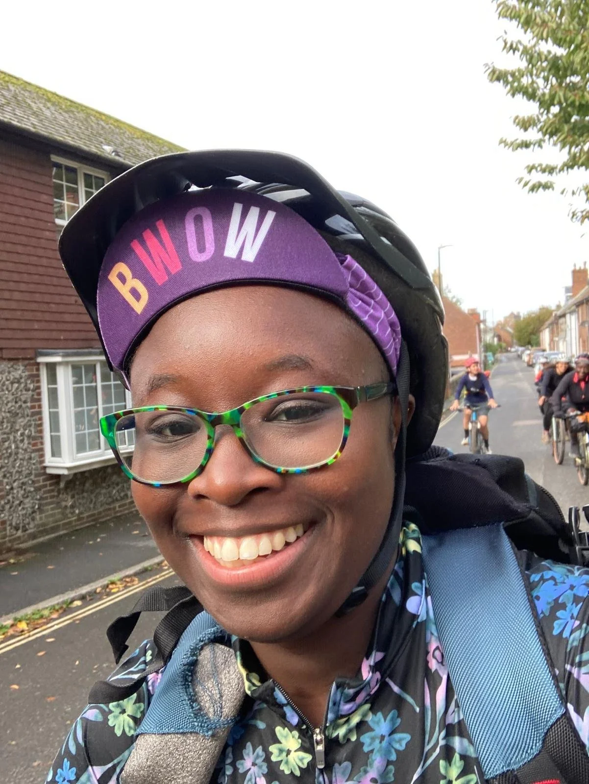 Smiling woman with glasses and a helmet, riding her bicycle on a street with other cyclists and residential houses in the background.