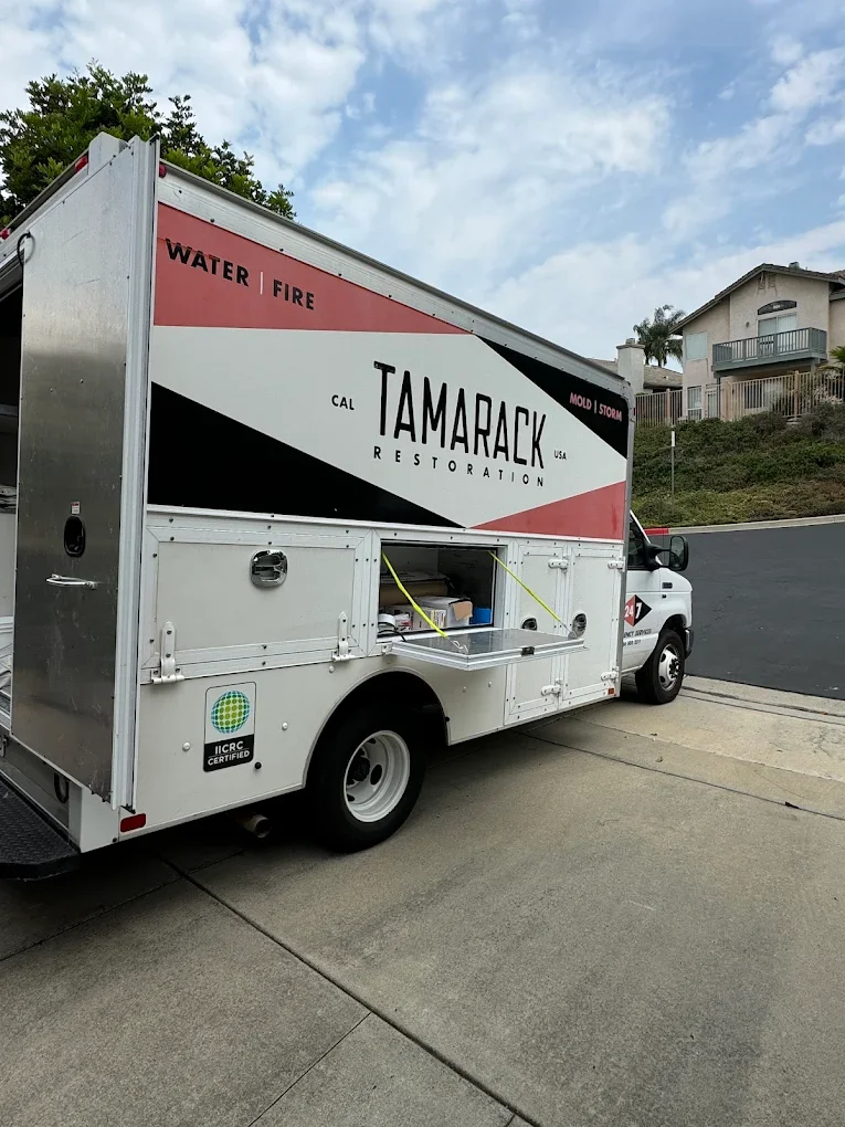 A Tamarack Restoration truck parked on a driveway in a residential neighborhood with houses and trees in the background.