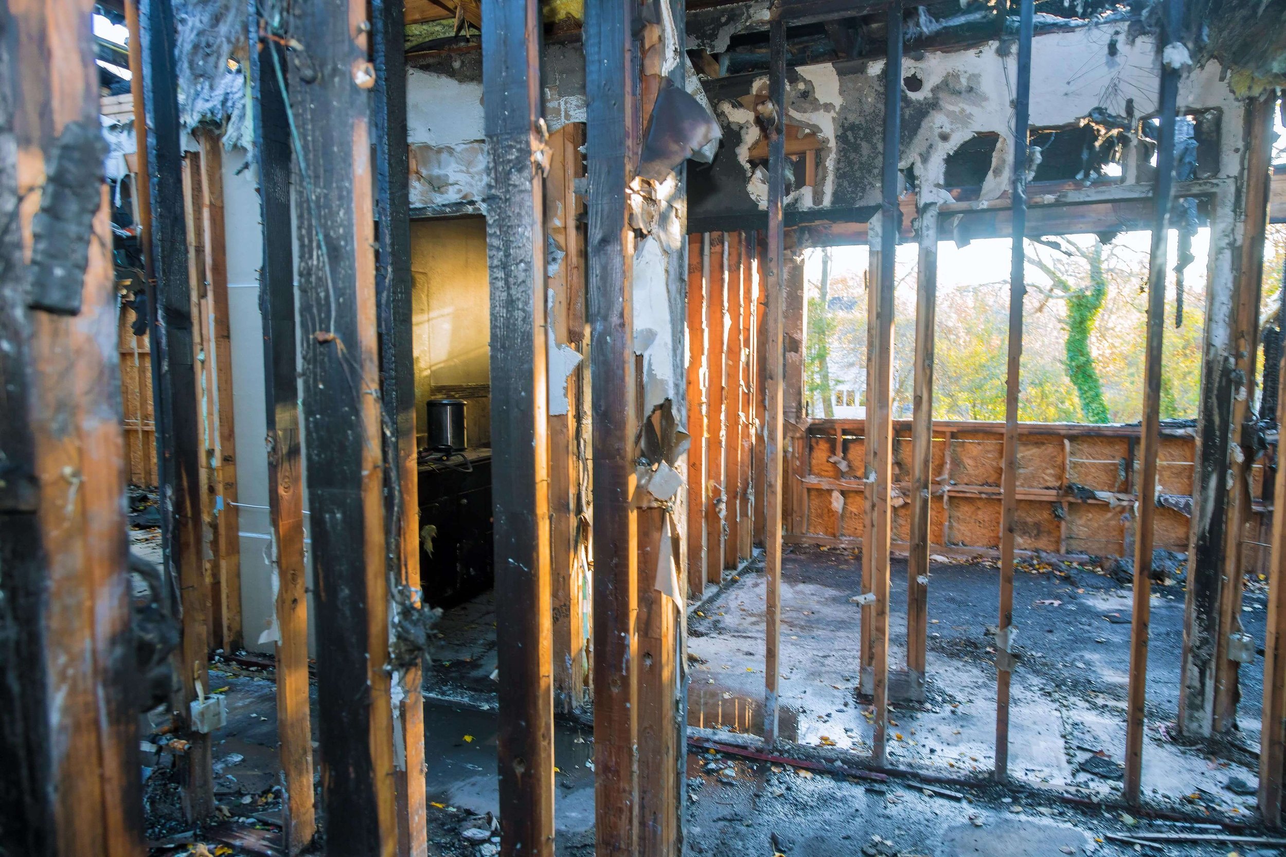 Interior of a house that has been burned, with charred wooden framing and walls, and debris on the floor. Through a damaged wall, a small portion of a kitchen with a microwave oven is visible.