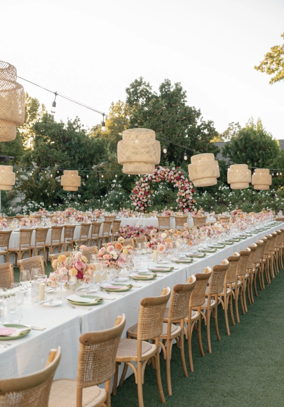 Long outdoor dining table decorated with pink and peach flowers, set with plates, glasses, and candles, under hanging wicker lanterns and string lights, with floral arch in the background.