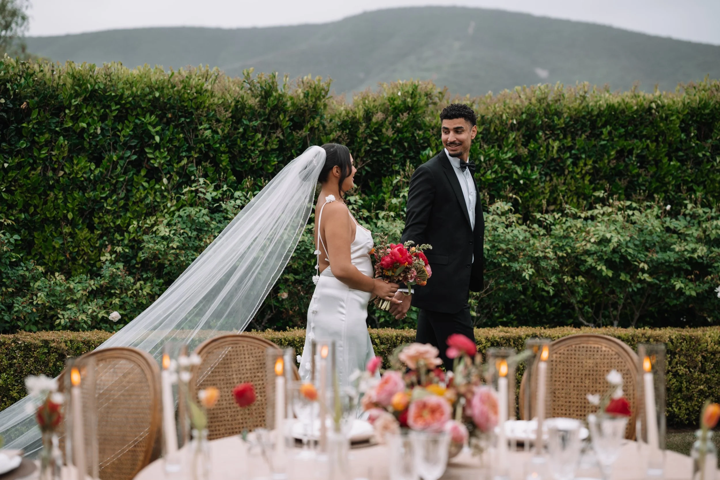 A bride and groom standing outdoors at their wedding reception, with the bride holding a bouquet of flowers and wearing a white dress with a veil, while the groom is dressed in a black tuxedo, in front of a lush green hedge and mountains in the backg