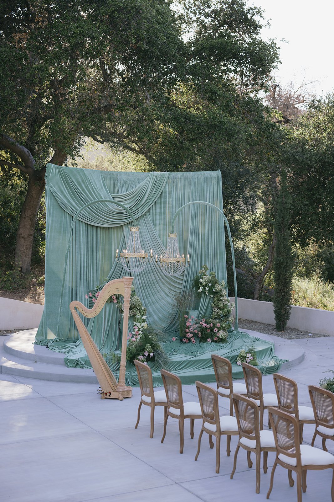 Outdoor wedding ceremony setup with a gold harp, lush floral arrangements, and elegant chandeliers on a pastel-colored backdrop with trees in the background.