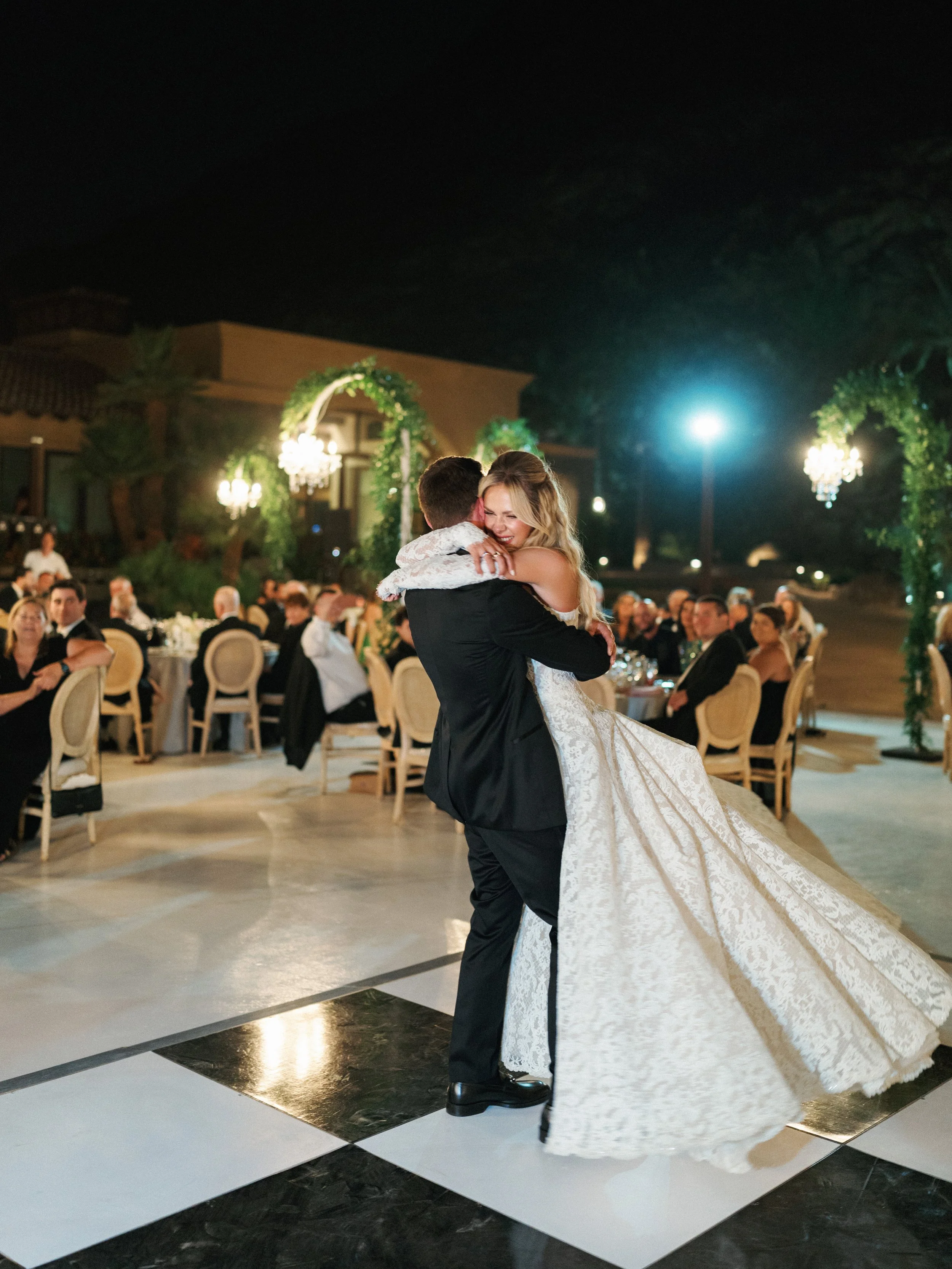 A bride and groom sharing their first dance at a wedding reception outdoors at night, with guests seated at tables around them, and festive lighting and greenery decoration.