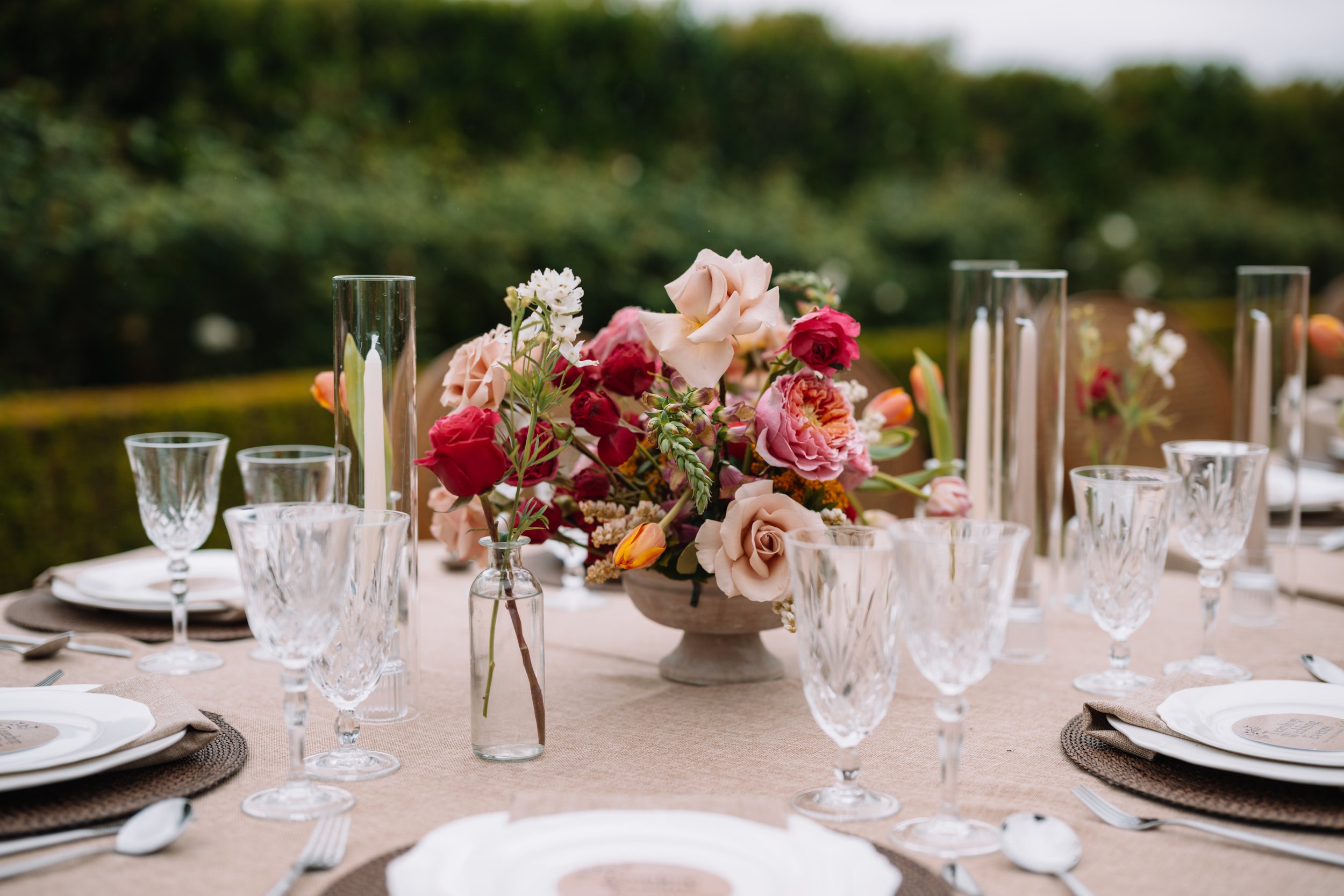 Elegant outdoor dining table decorated with a centerpiece of mixed pink, red, and peach flowers in a shallow stone bowl, surrounded by clear glassware, white plates, napkins, and silverware, set on a beige tablecloth with a blurred greenery backgroun