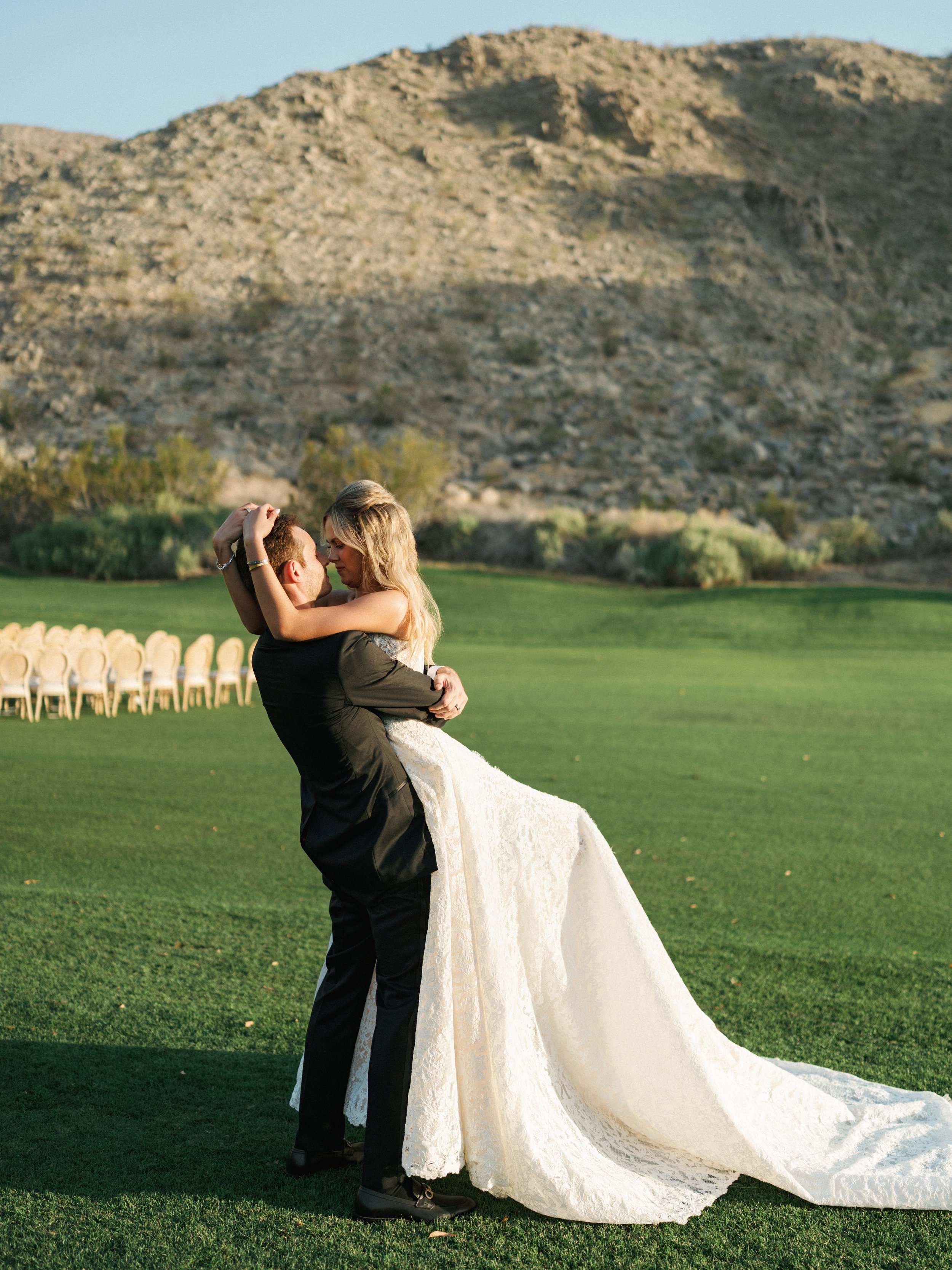 A newlywed couple dancing outdoors on a green field with mountains in the background during late afternoon or early evening.