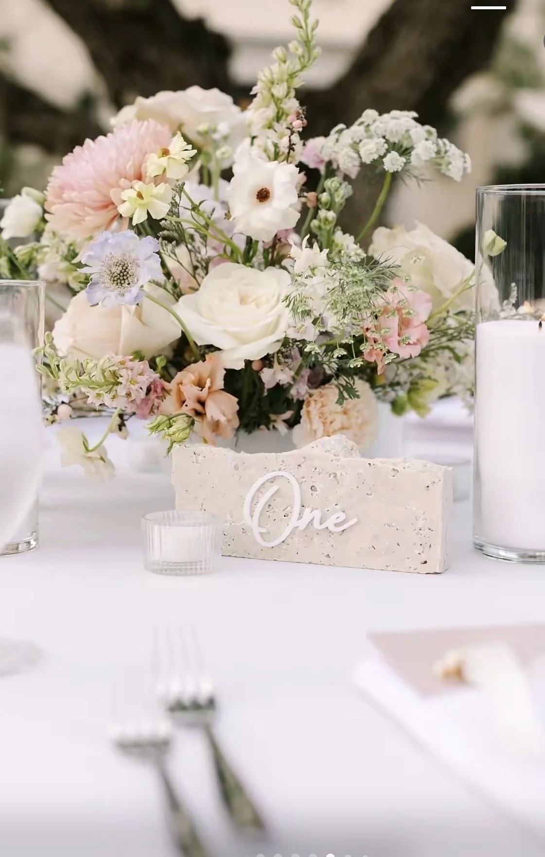 Elegant table centerpiece with pastel flowers, a 'One' table number, candles, and glassware for a wedding or celebration.
