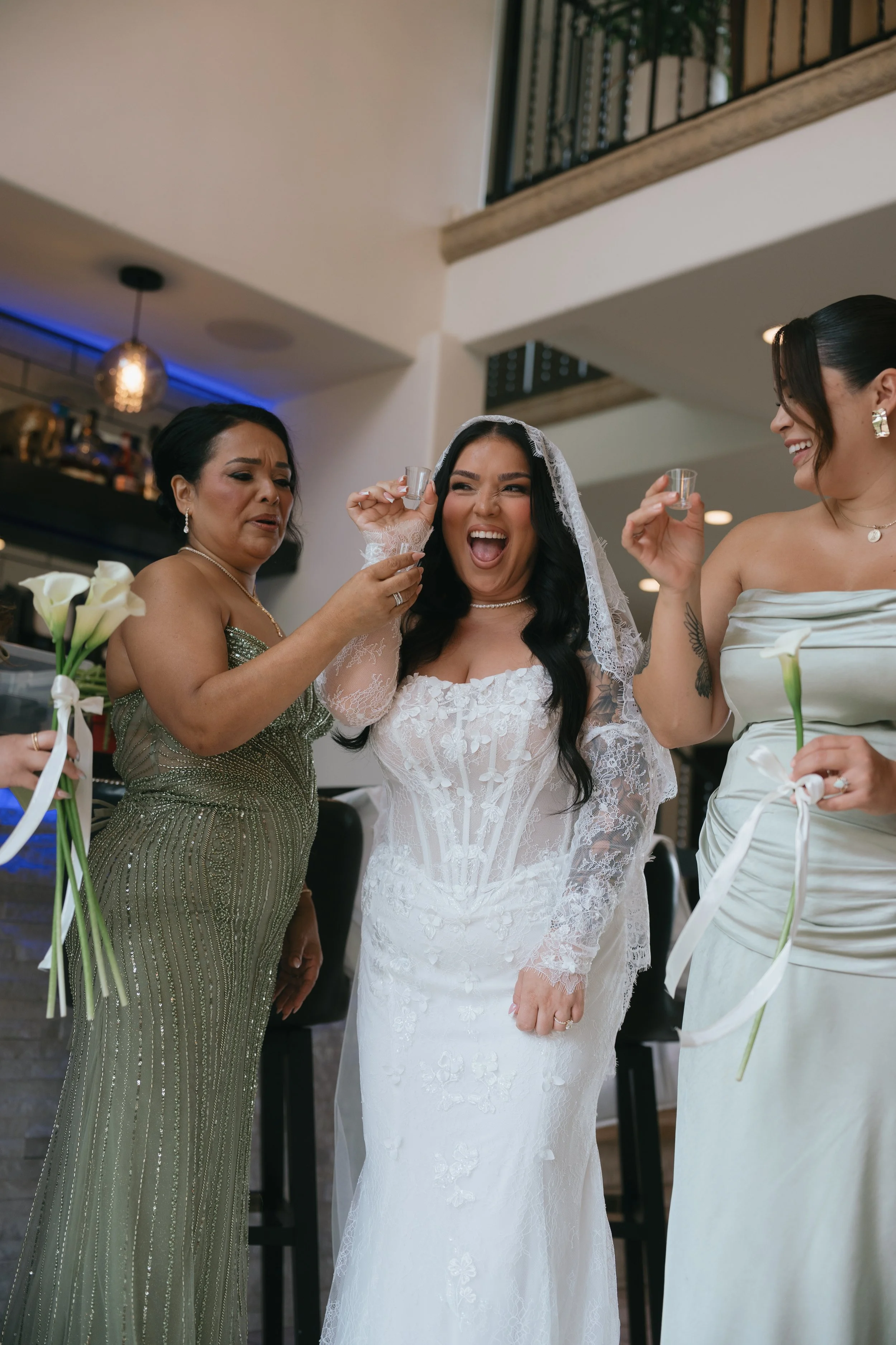 A bride and two women celebrating with shot glasses and calla lilies in a bright indoor setting.