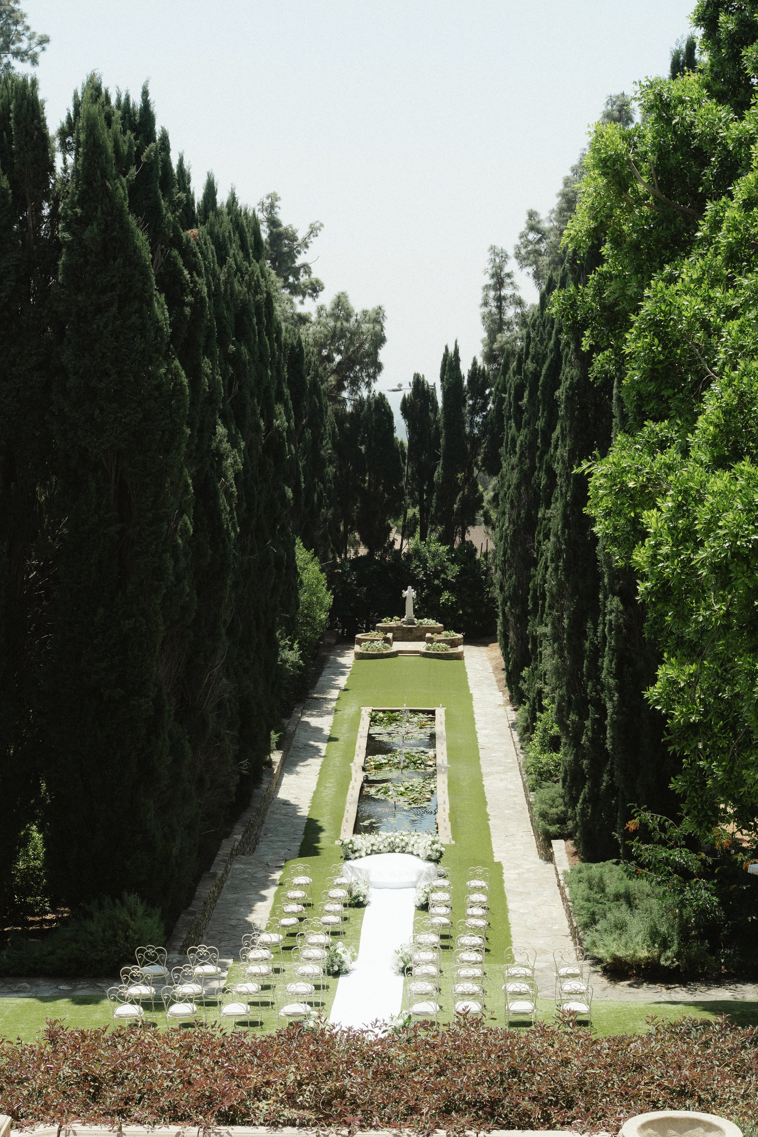 A garden with a long fountain, surrounded by tall trees and lined with white chairs for a wedding ceremony.