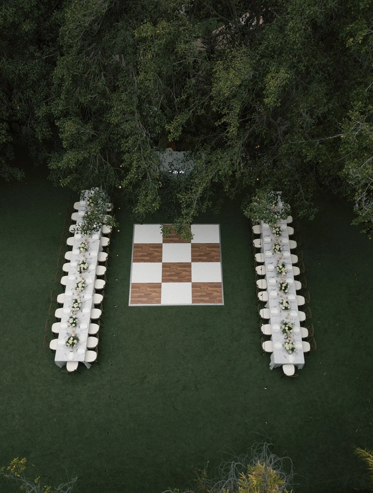 A top-down view of an outdoor event setup with two long tables on each side decorated with floral arrangements, positioned around a large outdoor chessboard on a green lawn, with trees in the background.