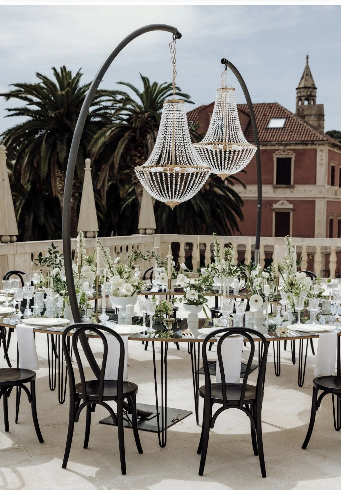 An outdoor event setup with black chairs around tables decorated with white flowers and glassware, under large chandeliers, with palm trees and historic buildings in the background.
