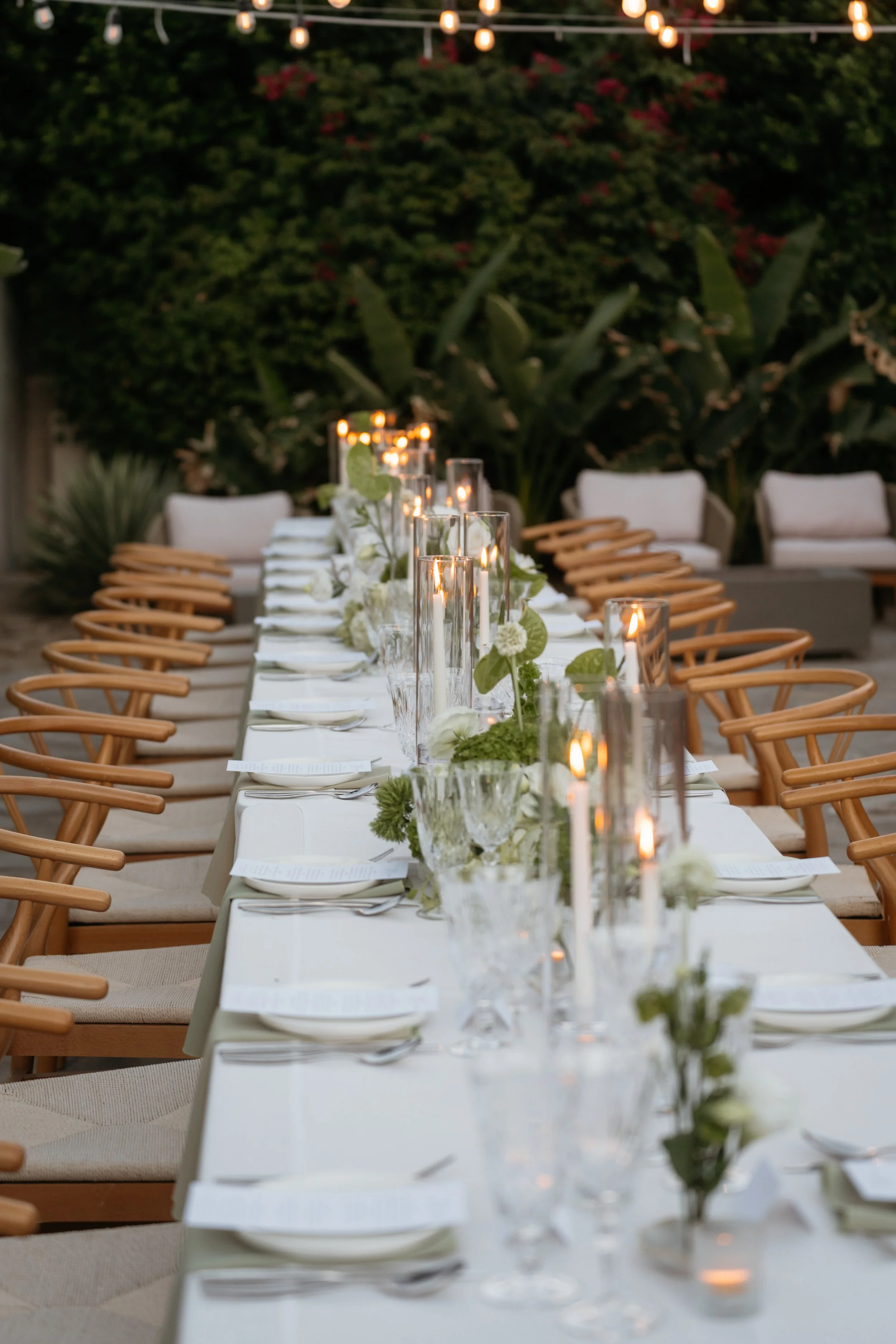 Elegant outdoor dining setup with a long white table, glass candle holders with lit candles, and floral centerpieces, surrounded by wooden chairs, with greenery and string lights overhead.