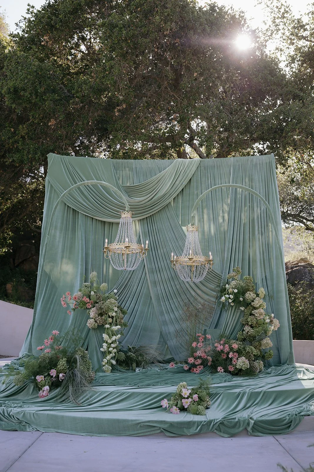 Elegant outdoor event setup with draped green fabric backdrop, two chandeliers, and floral arrangements of white and pink flowers. European-inspired wedding at Villa Vienna with elegant estate architecture