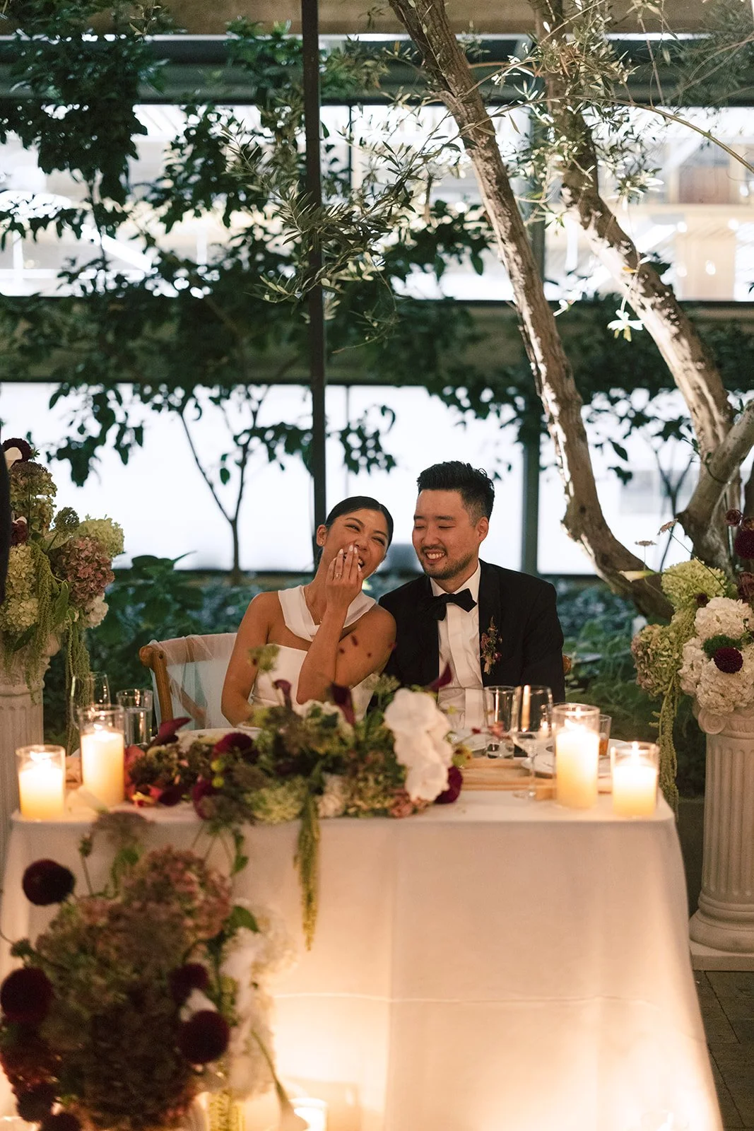A happy couple at their wedding reception, sitting at a decorated table with flowers and candles, outdoors with trees in the background.