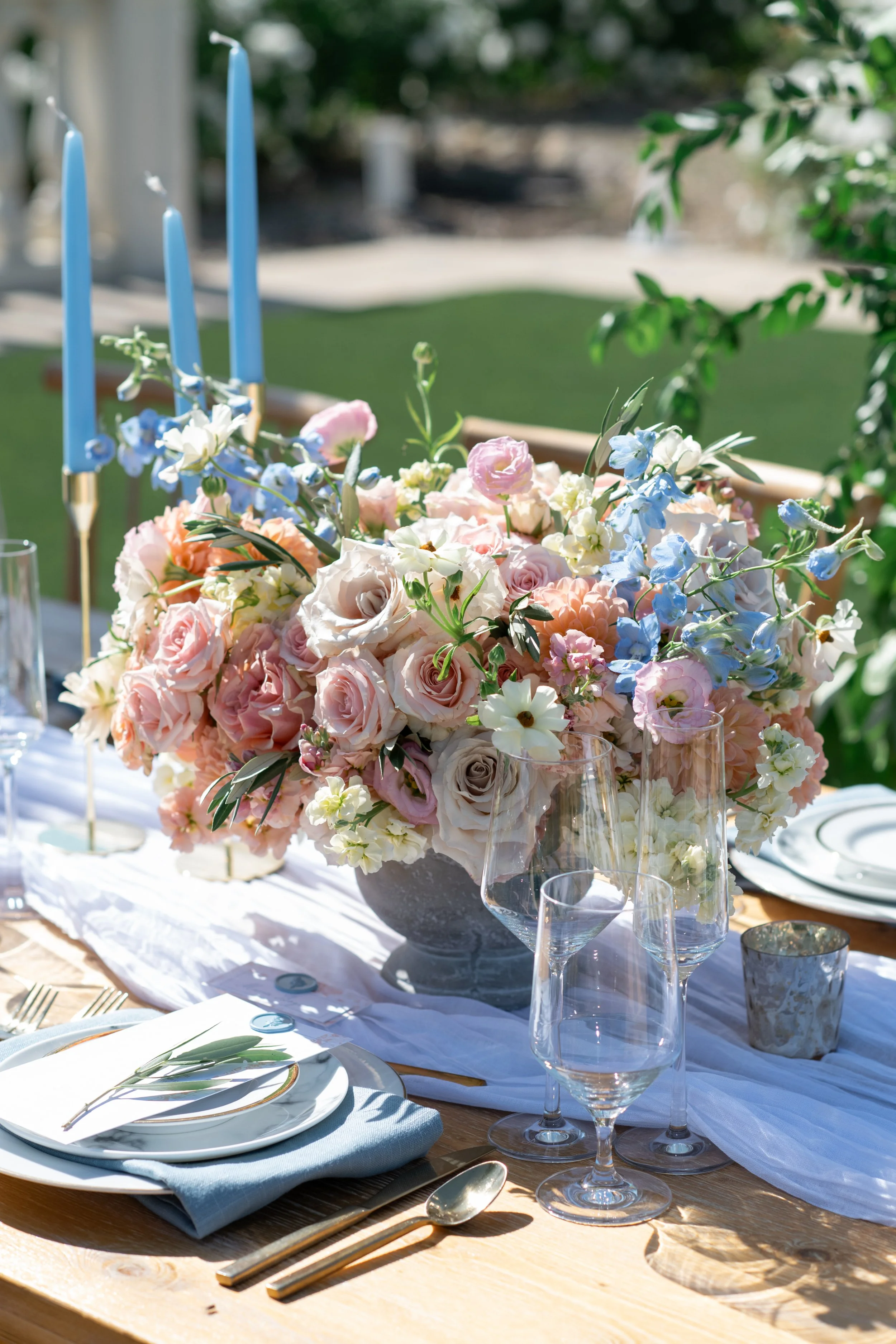 Elegant outdoor table setting with a large floral centerpiece of pink and white roses and other pastel flowers, surrounded by glasses, plates, cutlery, and blue candles, on a sunny day.