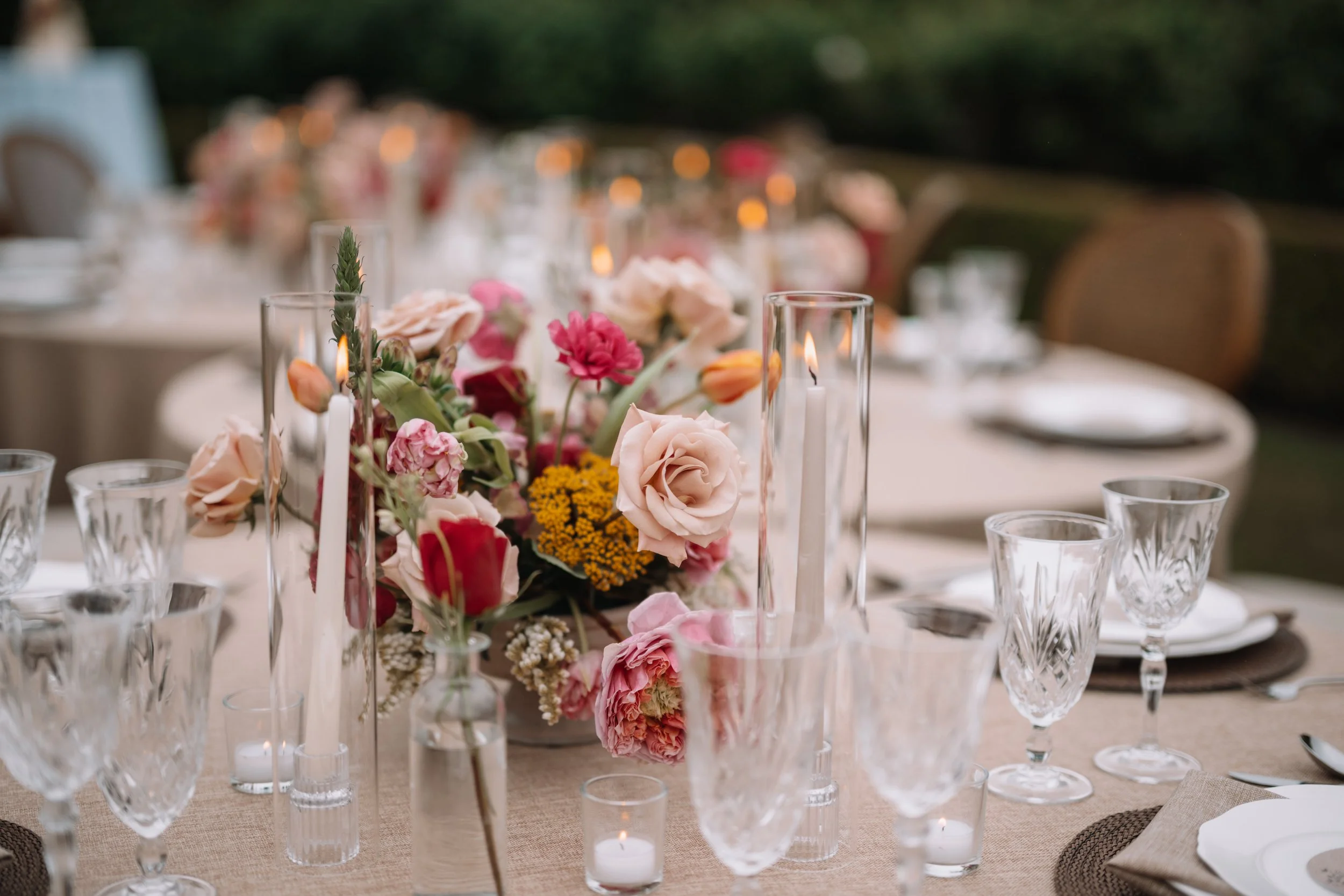 Elegant outdoor table set with floral centerpiece, candles, crystal glasses, and place settings for a celebration or wedding.