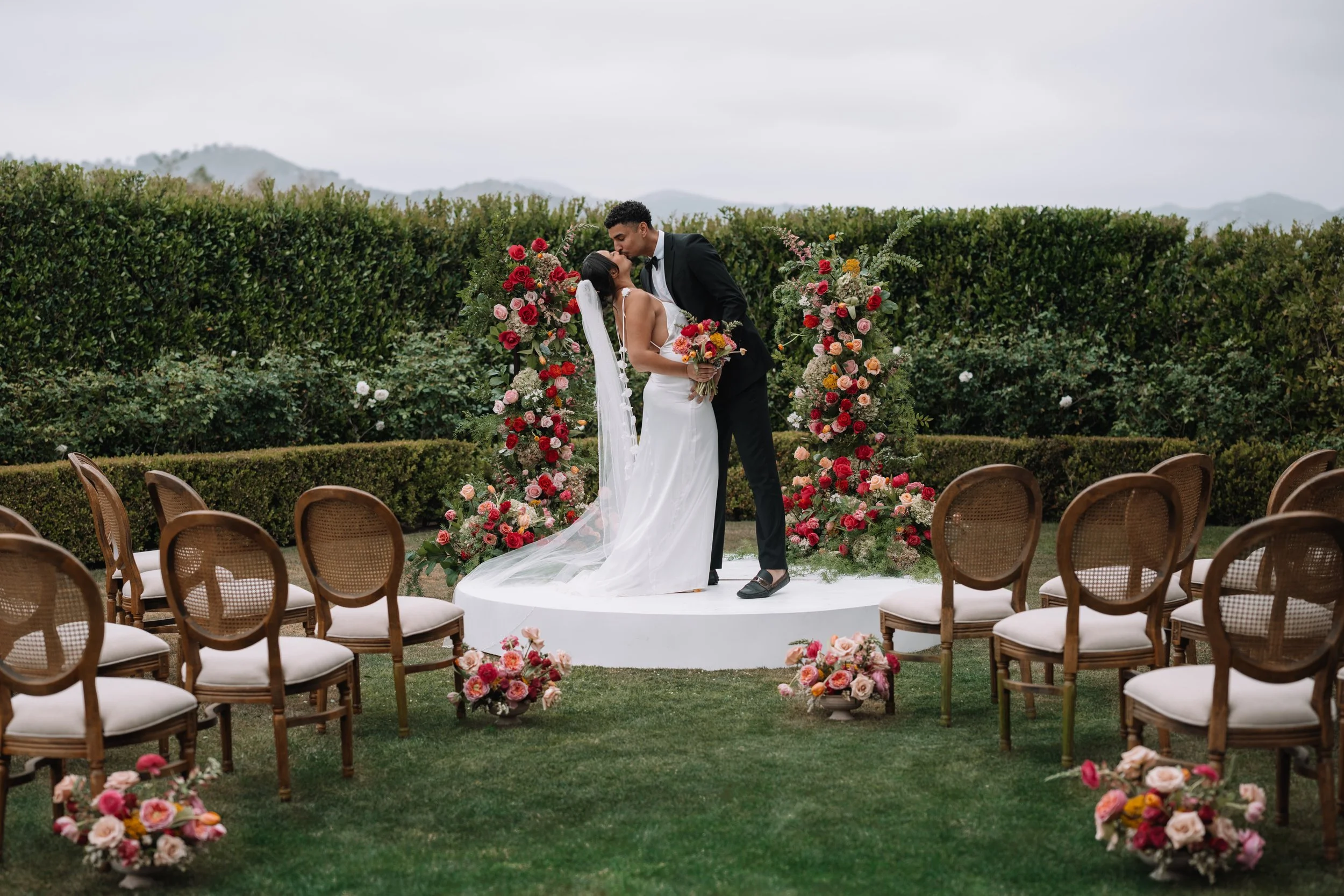 A couple kissing at their outdoor wedding ceremony, with the bride in a white wedding gown holding a bouquet, standing on a small stage surrounded by colorful floral arrangements. chairs are arranged for guests on the grass, with cloudy sky above and mountains in the background.