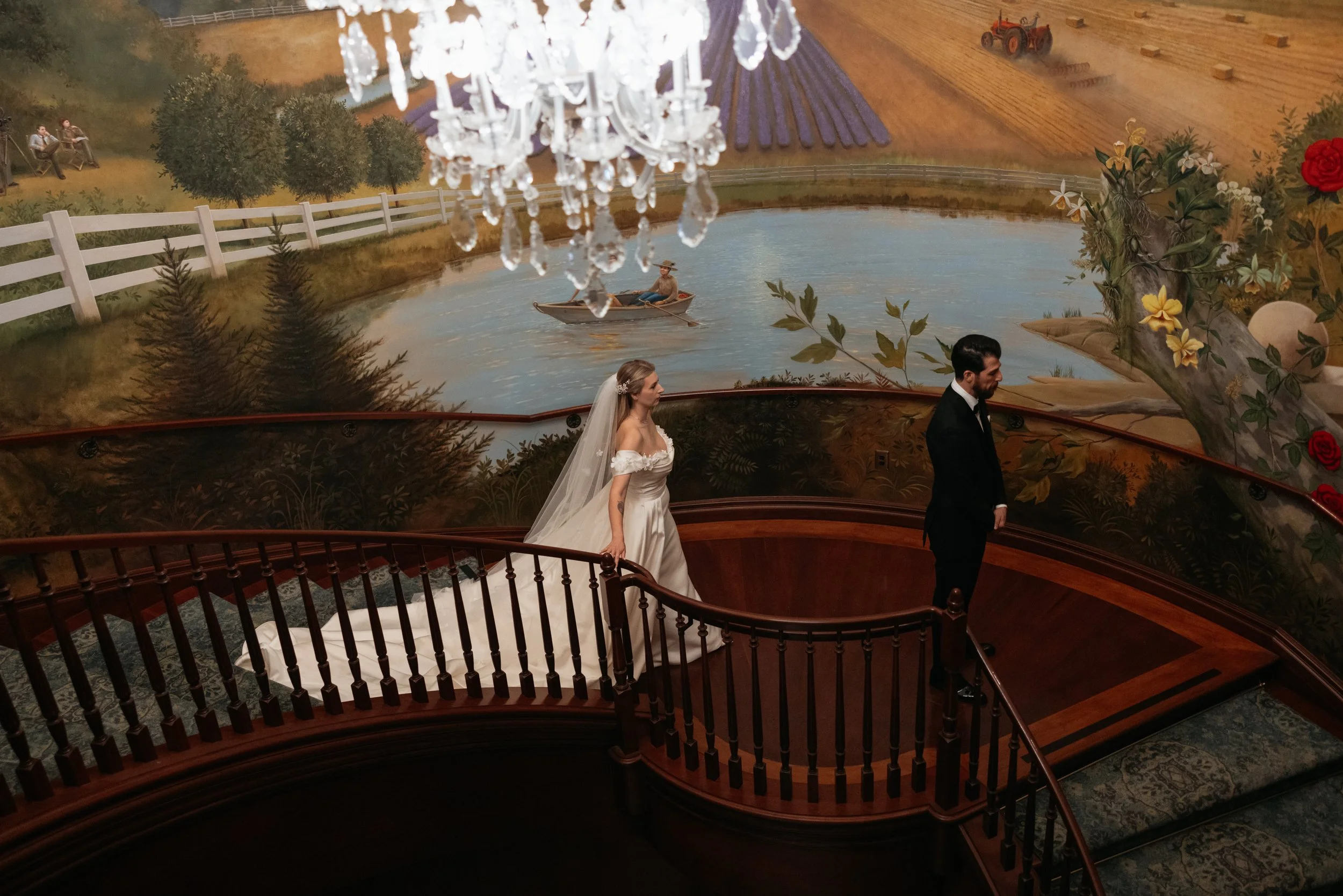 A bride and groom stand on a curved wooden balcony with a mural depicting a lakeside scene with trees, a boat, and a tractor in the background. The bride wears a white wedding gown and veil, while the groom is in a black suit. A crystal chandelier ha