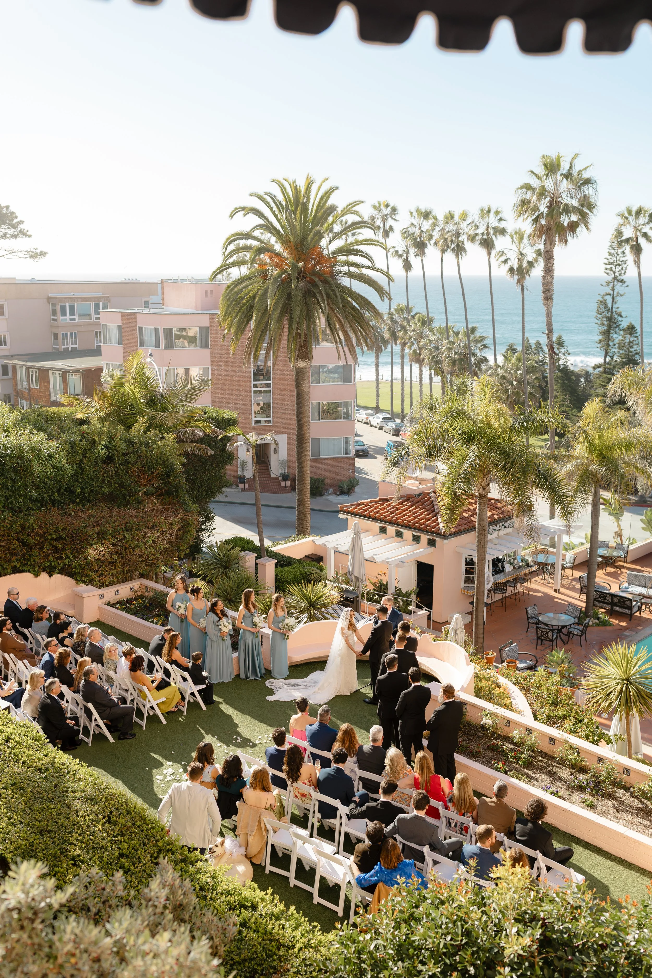 Wedding ceremony outdoors at a venue with palm trees, ocean view, and guests seated in white chairs.