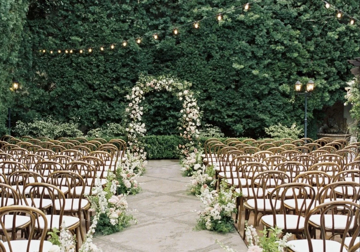 Outdoor wedding ceremony setup with rows of wooden chairs on each side of a stone aisle, decorated with floral arrangements, leading to a floral arch against a background of green trees and string lights overhead.