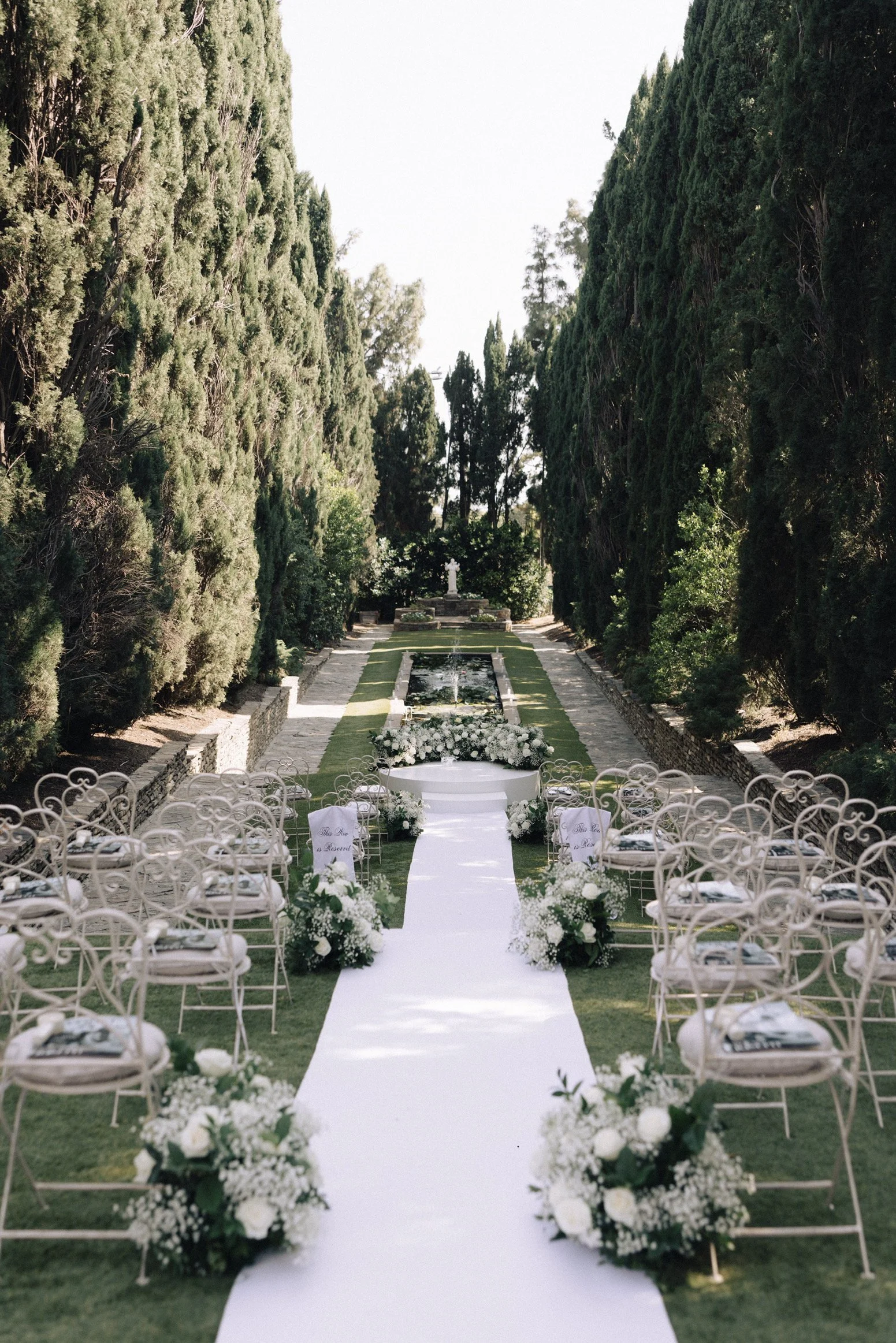 Outdoor wedding ceremony with white aisle runner, surrounded by chairs with white covers and flower arrangements, flanked by tall green trees, leading to a small altar with a cross.