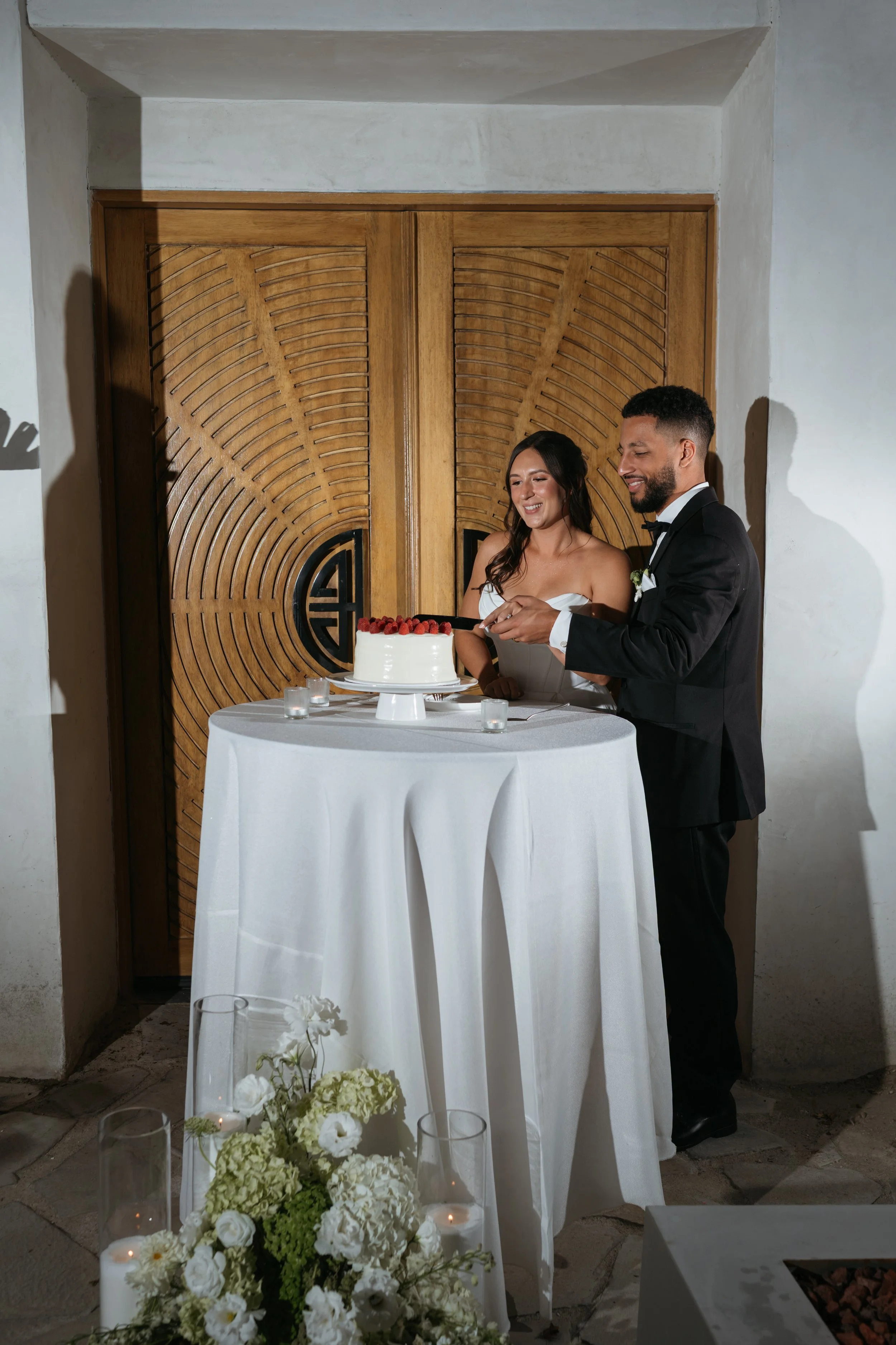 A bride and groom cutting a wedding cake at their reception, standing near a white-covered table with candles and floral arrangements.