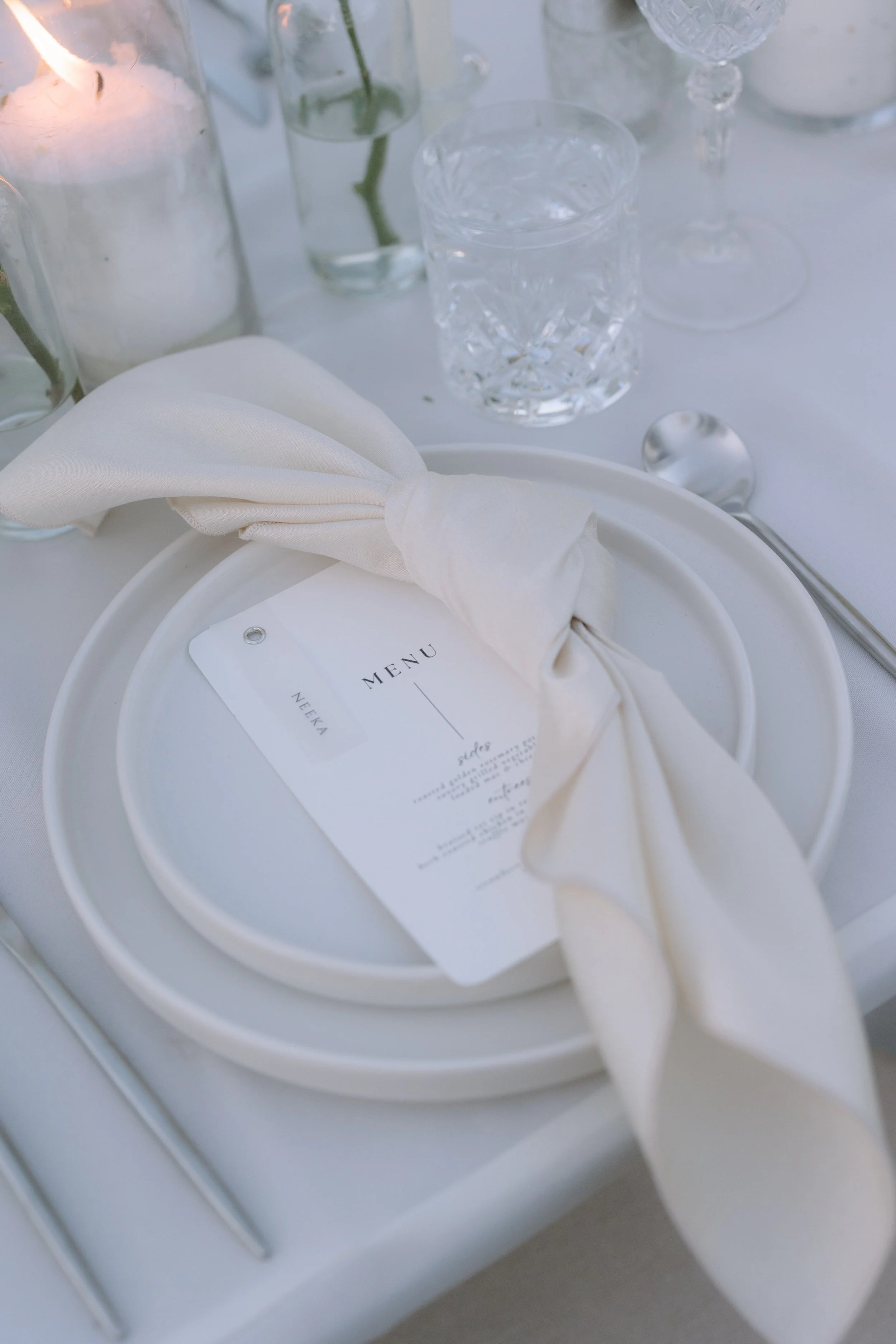 Elegant table setting with white plates, a cream-colored napkin, a menu card, water glasses, and a candle in a glass holder.