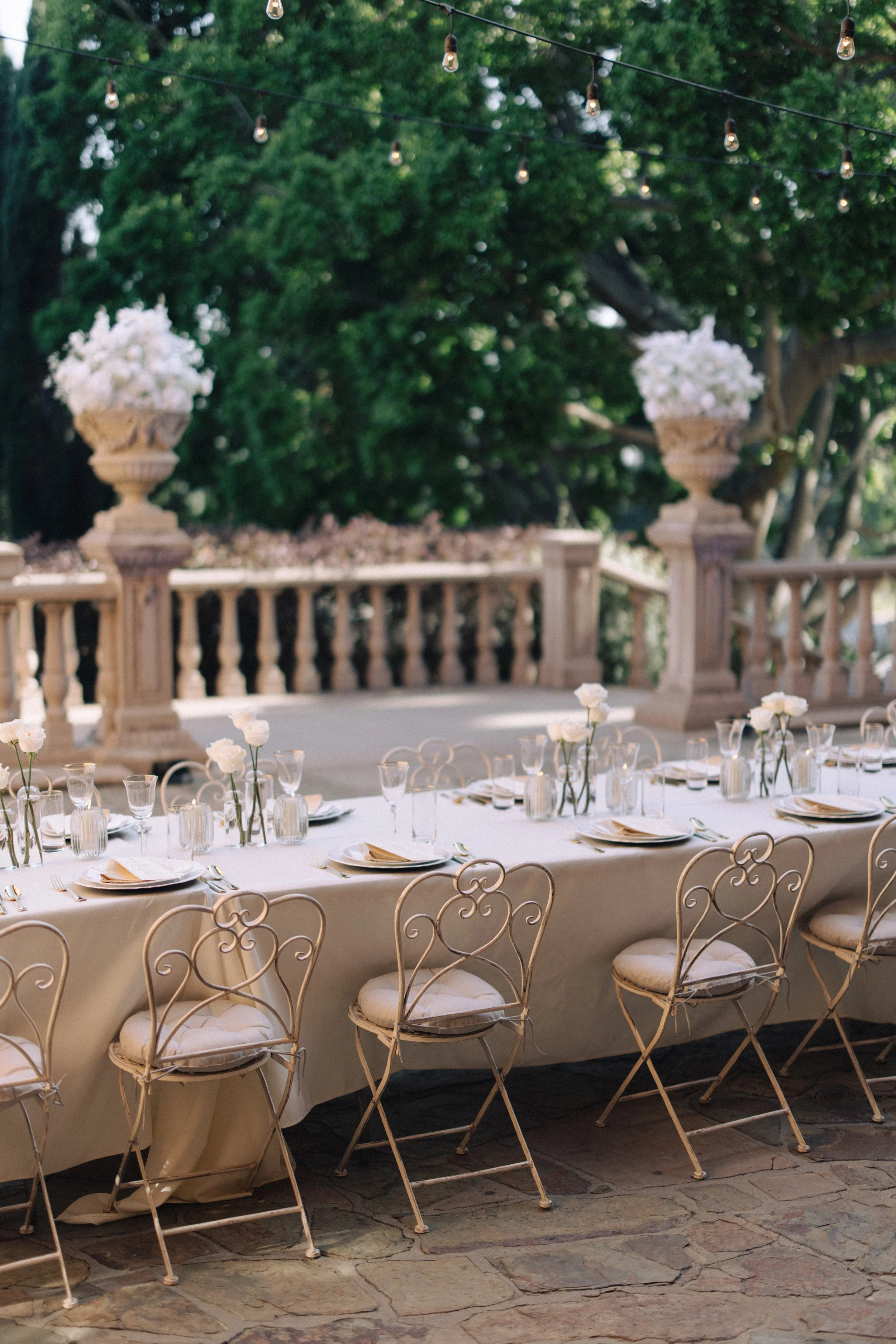 Outdoor event setup with a long dining table, elegant chairs, white tablecloth, floral centerpieces, and string lights hung overhead on a patio or garden area, with trees in the background.
