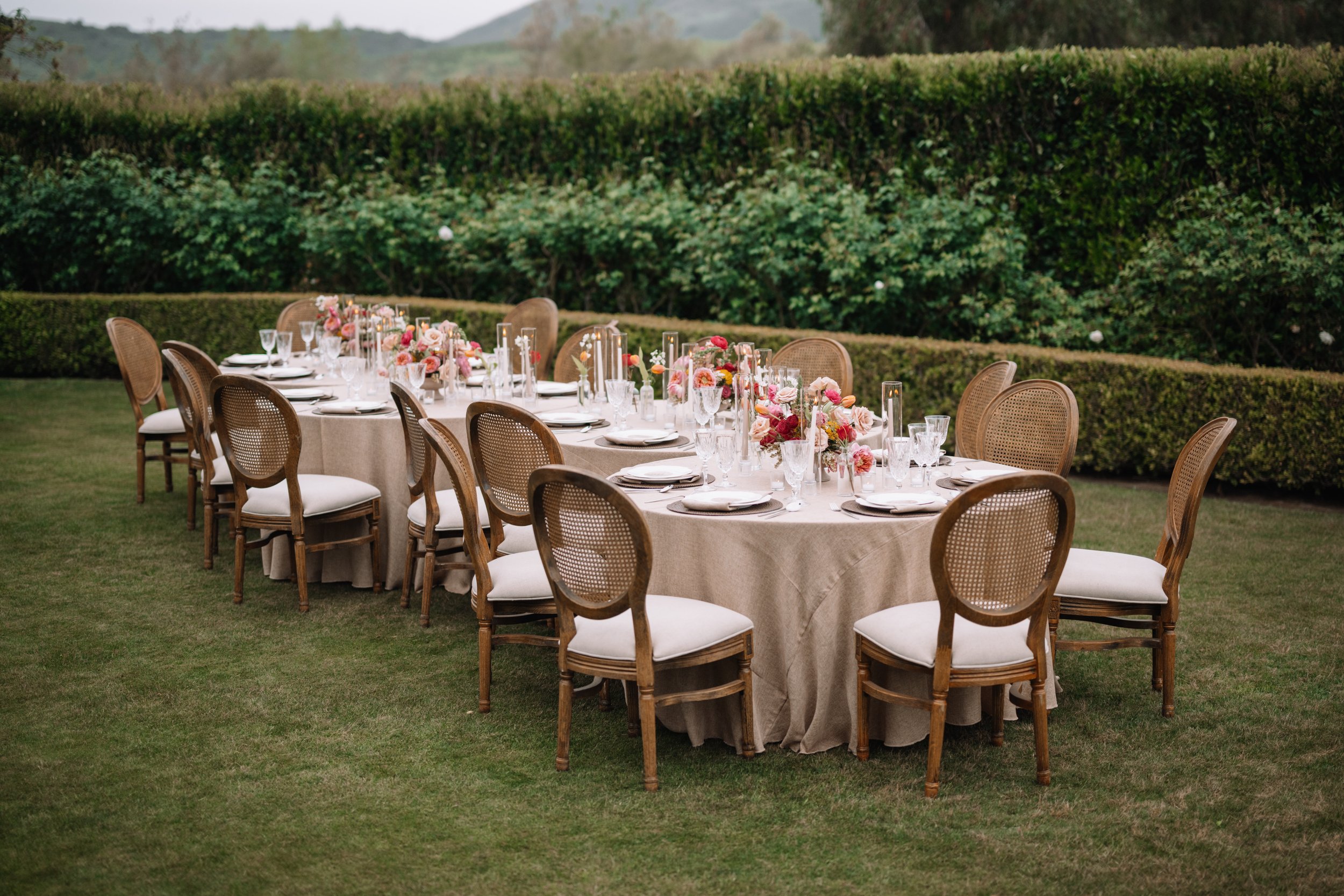 Long outdoor dining table with floral centerpieces, set with plates, glasses, and napkins, surrounded by wooden chairs with white cushions, on a grassy lawn with hedges and hills in the background.